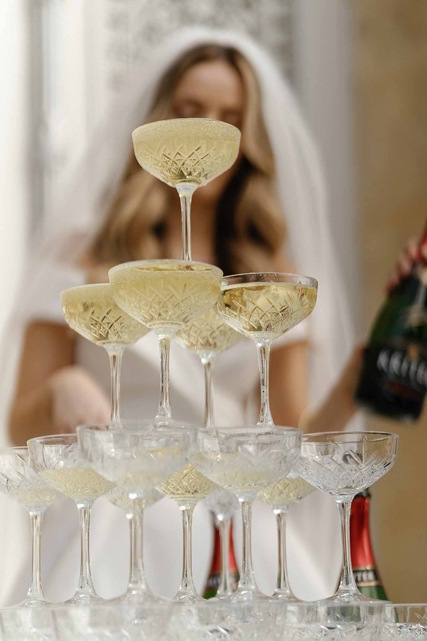 A close-up detail shot of a champagne tower built from crystal-cut coupe glasses filled with pale golden sparkling wine, arranged in a classic pyramid formation across at least three visible tiers. In the soft-focus background, a bride wearing a white veil and white dress is pouring champagne from a dark green bottle into the tower. A second champagne bottle with a red foil cap is visible at the base of the tower in the foreground. The setting appears to be indoors with neutral warm tones, and the overall decor style is classic and formal.