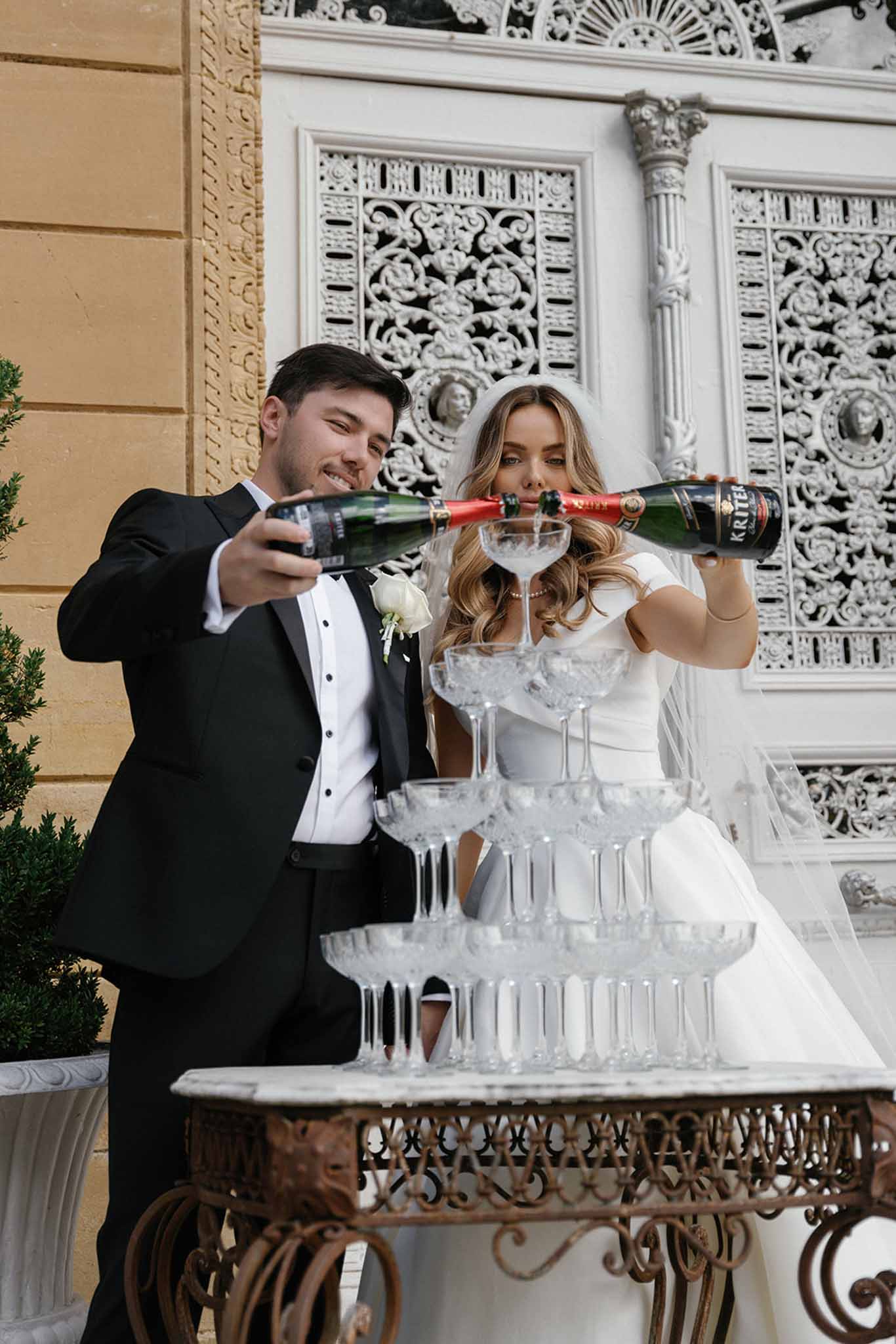 The bride and groom are pouring Kriter sparkling wine into a champagne tower of crystal coupe glasses stacked on an ornate wrought-iron table, photographed outdoors against the facade of a grand classical building with white decorative ironwork panels and carved stone detailing. The groom wears a black tuxedo with a white rose boutonniere, while the bride wears a white short-sleeve fitted gown with a long veil and loose wavy blonde hair. The champagne tower consists of approximately five tiers of coupe glasses, with champagne visibly cascading through the top glasses. The shot is a mid-range portrait taken from a low angle looking slightly upward, placing the couple and the ornate architectural backdrop prominently in the frame.