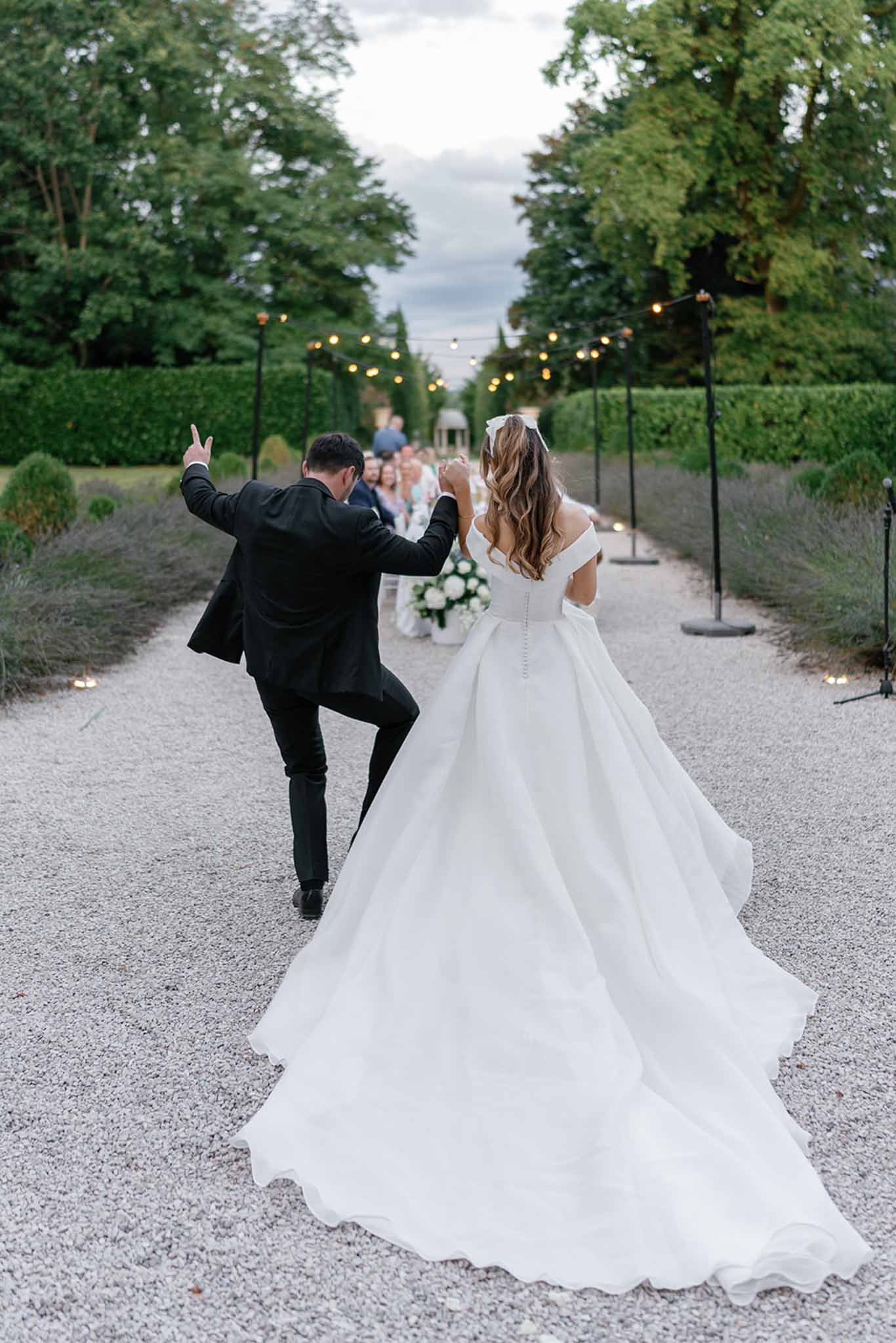 The bride and groom are photographed from behind as they walk along a formal gravel garden alley toward a group of seated guests in the distance, appearing to make a celebratory entrance. The groom, wearing a black suit, has one arm raised with a playful gesture while the other hand holds the bride's. The bride wears an off-the-shoulder ivory ball gown with a corseted back, button-down train, and a white bow hair accessory in her long wavy hair. The outdoor setting features a manicured formal garden with trimmed hedgerows, topiary, and lavender borders lining the path, strung with warm Edison bulb festoon lights on metal poles. Small candles are placed along the path edges, and a white floral arrangement is visible among the guest seating in the background. The composition is a wide portrait shot taken at dusk, capturing the full sweep of the gown's train across the gravel.