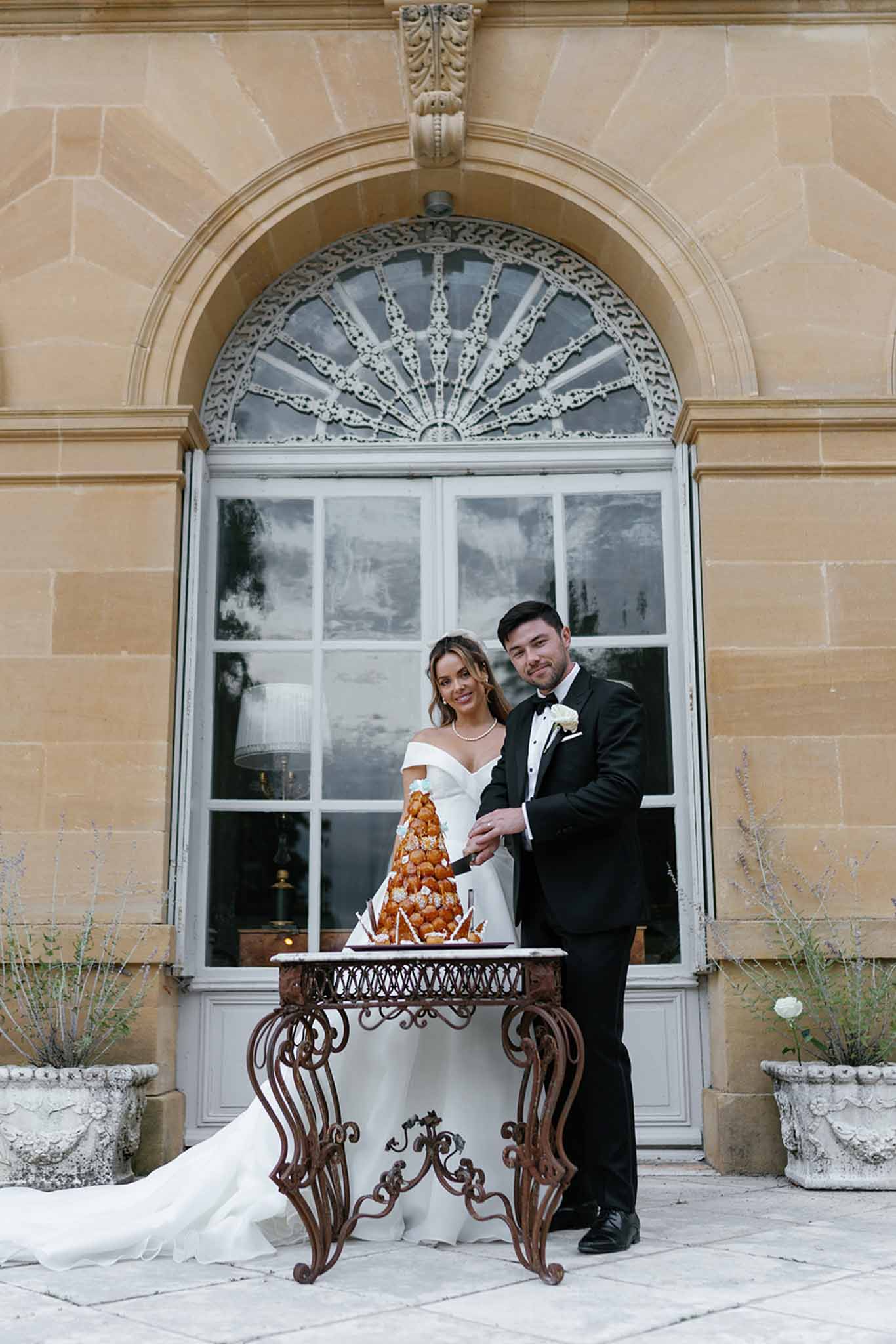 The bride and groom are cutting a croquembouche together on an ornate wrought-iron table positioned on an outdoor stone terrace in front of a chateau facade. The croquembouche is a tall cream puff tower decorated with spun sugar and dusted with powdered sugar. The bride wears an off-the-shoulder white gown with a cathedral-length train, paired with a pearl necklace, while the groom wears a classic black tuxedo with a white boutonniere and bow tie. The setting features a large arched window with an elaborate fan-shaped iron transom above it, flanked by two stone urns with plantings. The overall styling is classic and formal, and the shot is a medium portrait framing both figures full-length against the chateau architecture. Potential venue feature image.