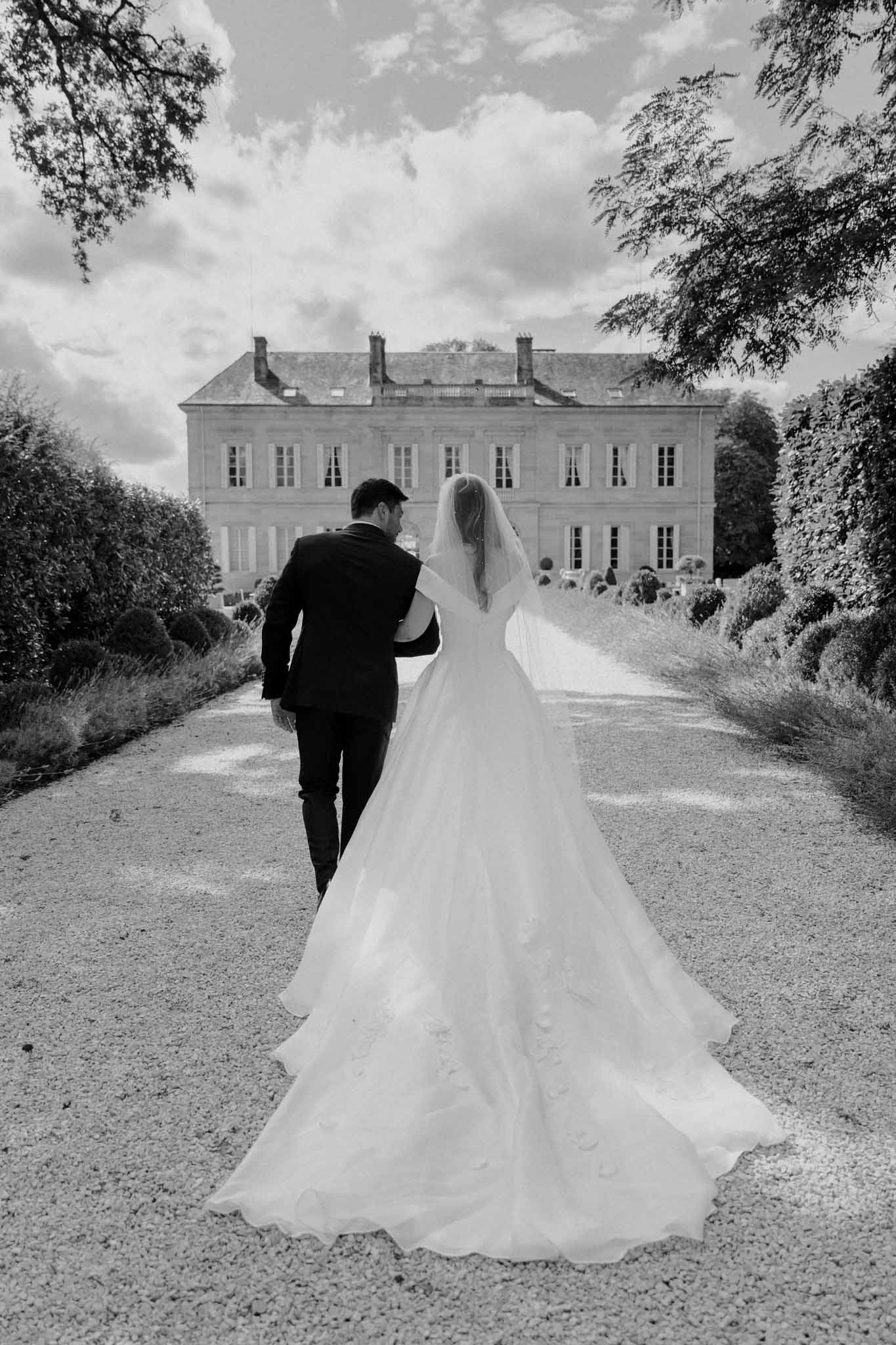 Black-and-white couple portrait taken outdoors on a gravel path leading directly toward a large French château with classical 18th-century architecture, symmetrical façade, and tall shuttered windows. The bride and groom walk away from the camera arm-in-arm, their faces turned slightly toward each other; the bride wears a full ball-gown style dress with an extended cathedral-length train spreading across the path, delicate appliqué detailing visible on the skirt, and a long flowing veil, while the groom wears a dark suit. The image is shot in high contrast black and white with deep shadows in the foreground and bright mid-tones on the château façade. The composition is a wide rear-facing portrait with the couple centered on the path, creating a strong leading-line perspective toward the venue. Potential venue feature image.