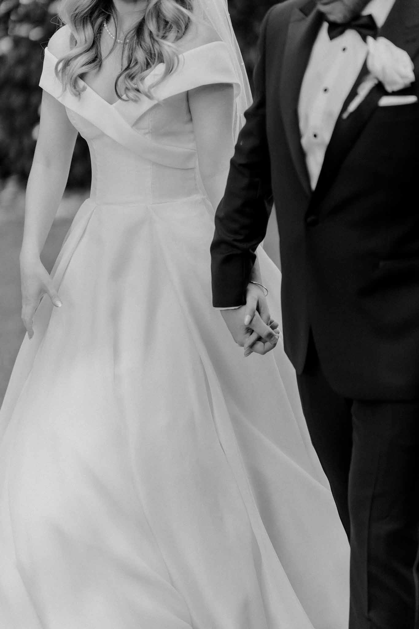 This is a black-and-white portrait of a bride and groom holding hands while walking outdoors, cropped from approximately the shoulders to mid-thigh. The bride wears a structured off-the-shoulder ball gown with a fitted bodice and a full, flowing skirt with a lightweight, billowing quality, along with a veil and a delicate pearl necklace; her wavy hair falls over one shoulder. The groom is dressed in a classic black tuxedo with a bow tie and a white boutonniere on his lapel. The image is close-up and detail-focused, emphasizing the couple's joined hands and the contrast between the bright white of the gown and the deep black of the tuxedo, with soft, even tones throughout.
