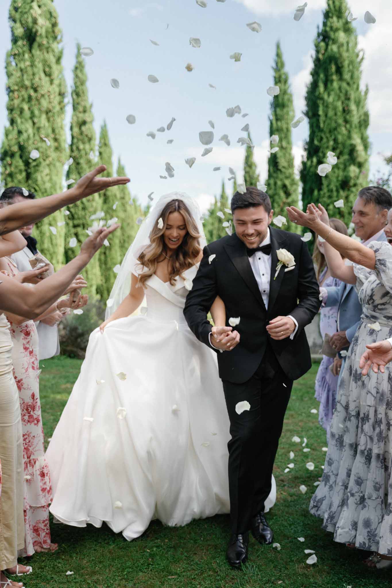 The bride and groom walk through a petal toss exit during an outdoor ceremony recessional, with approximately a dozen guests lining both sides throwing white rose petals into the air. The setting is an outdoor garden or estate grounds flanked by tall cypress trees. The bride wears an off-shoulder white ball gown with a long veil and has loose wavy hair, while the groom wears a black tuxedo with satin lapels, a black bow tie, and a white rose boutonniere. Guests are dressed in a mix of floral-print and pastel outfits. The shot is a mid-range portrait taken straight-on, capturing the couple laughing and holding hands as petals fall around them.