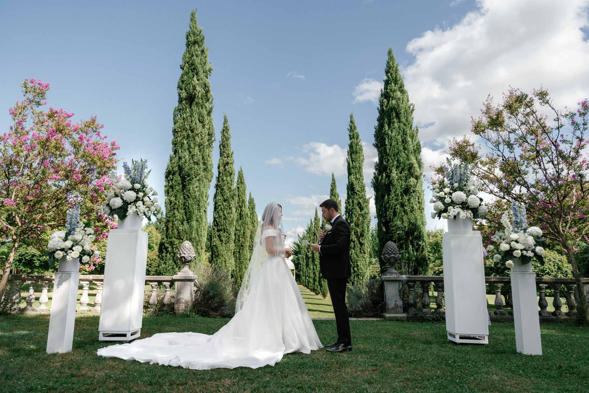 Bride and groom exchanging vows during outdoor ceremony in formal garden with cypress trees and floral arrangements