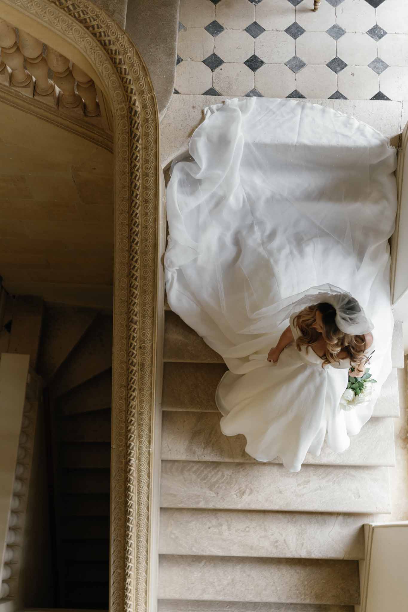 Bride in ivory gown descending stone staircase in classical interior with ornate architectural details