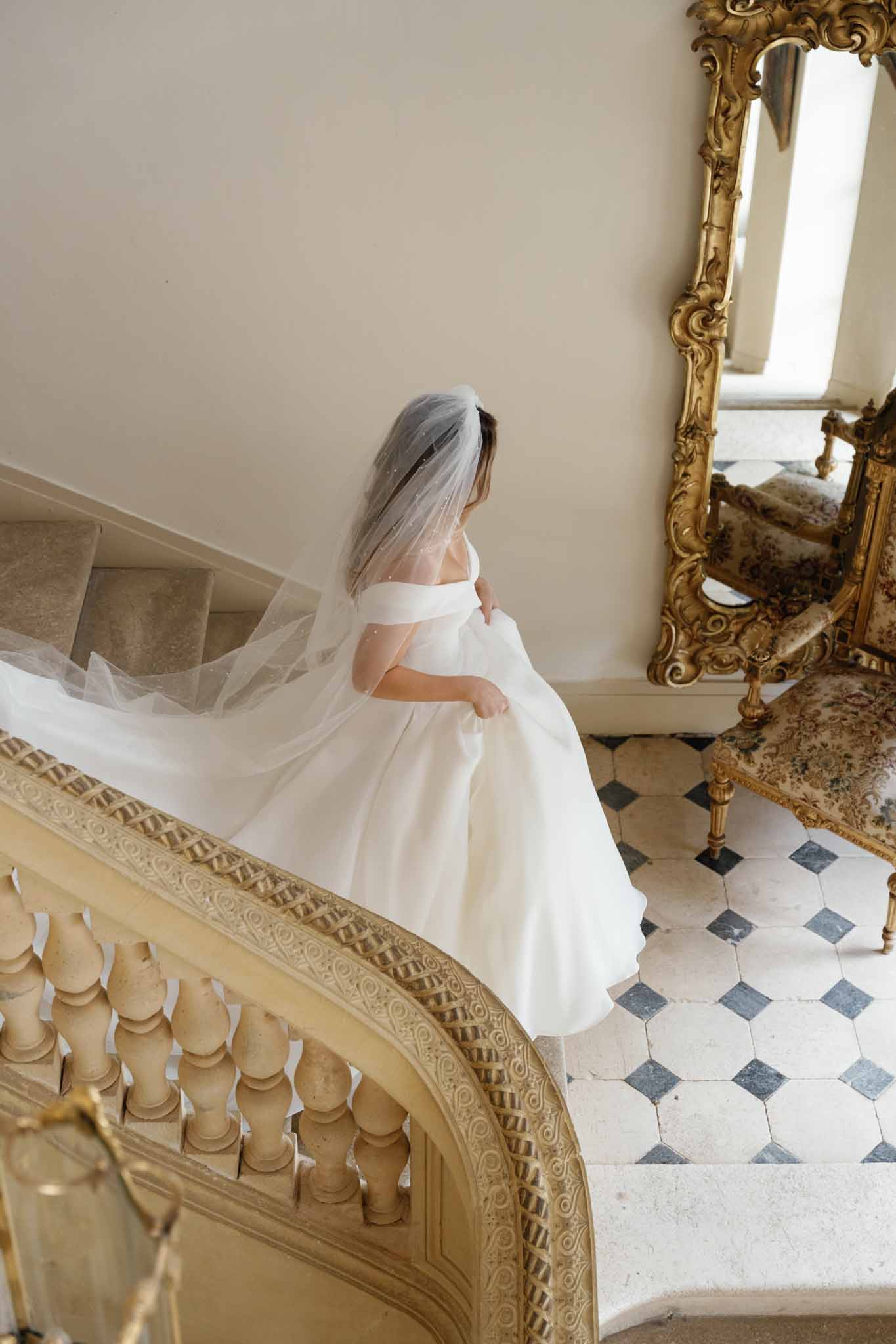 Bride in ivory off-shoulder dress and tulle veil in elegant classical interior hallway with ornate mirror