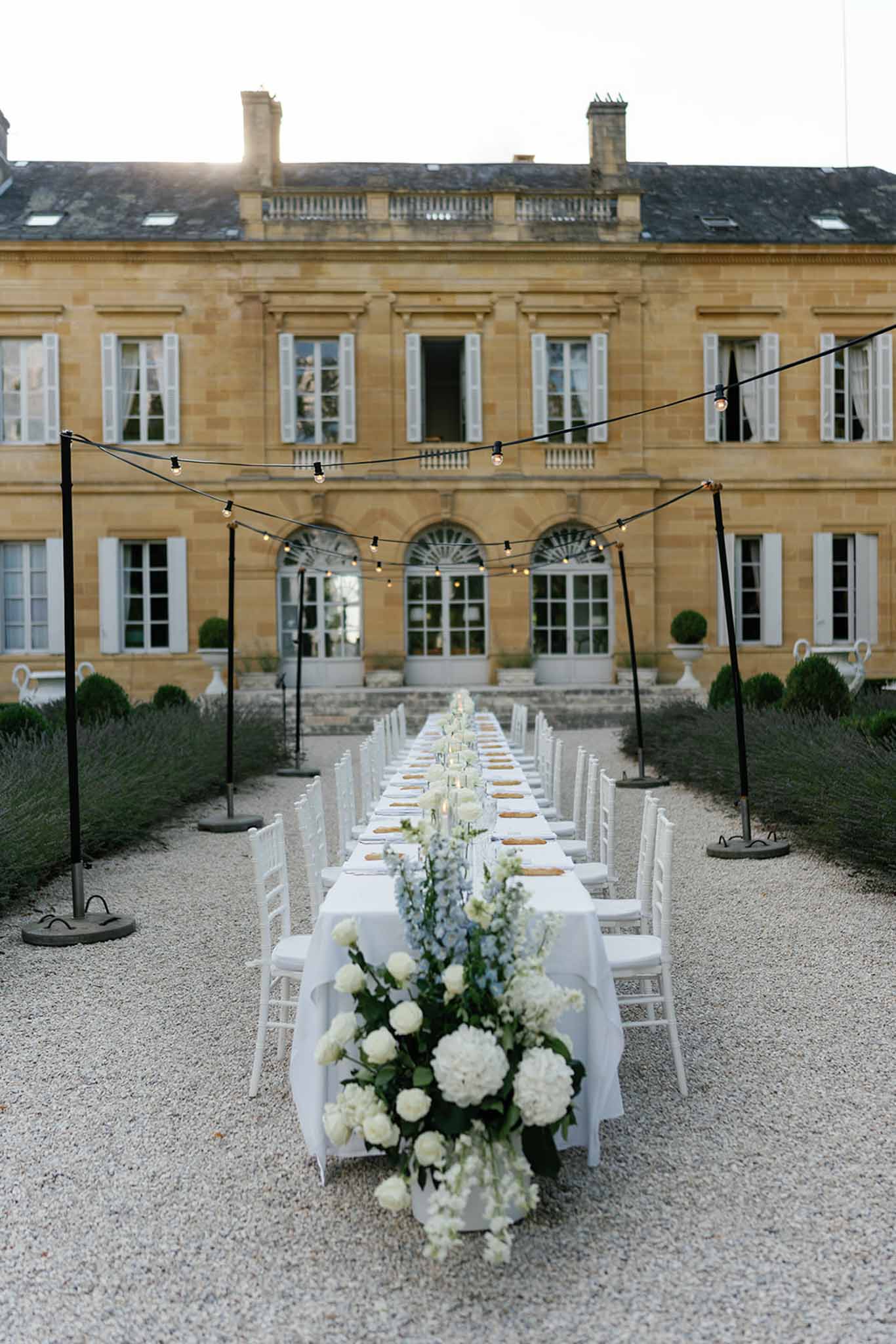 An outdoor wedding reception dinner is set up on the gravel forecourt of a large French chateau, with a single long banquet table running directly toward the building's entrance. The table is dressed in a white linen tablecloth and lined with white chiavari chairs on both sides, seating approximately 20–24 guests. A lush floral runner extends the full length of the table, composed of white hydrangeas, white peonies, pale blue delphiniums, and dark green foliage, with candles and gold charger plates visible along the place settings. Edison-bulb string lights are strung overhead on black poles above the table, providing warm ambient lighting as dusk approaches. The overall decor palette is white, soft blue, and gold against the warm honey-toned stone of the chateau facade, reflecting a classic and refined outdoor dining aesthetic. Wide shot taken from ground level behind one end of the table, framing the chateau as backdrop. Potential venue feature image.