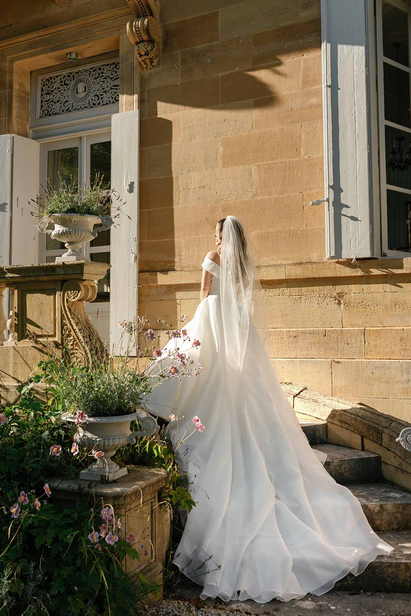 A bridal portrait taken outdoors at a French chateau, showing a single bride standing on stone steps alongside the building's facade. The bride wears an off-the-shoulder white ball gown with a full, flowing skirt and a long cathedral-length veil, and she is turned slightly away from the camera looking to one side. The chateau exterior features warm golden limestone masonry, white-painted shutters, ornate ironwork transom panels, and white stone pedestal urns planted with greenery and small pink cosmos-style flowers. The shot is a full-length portrait framed vertically, with strong directional sunlight casting defined shadows across the stone facade. Potential venue feature image.