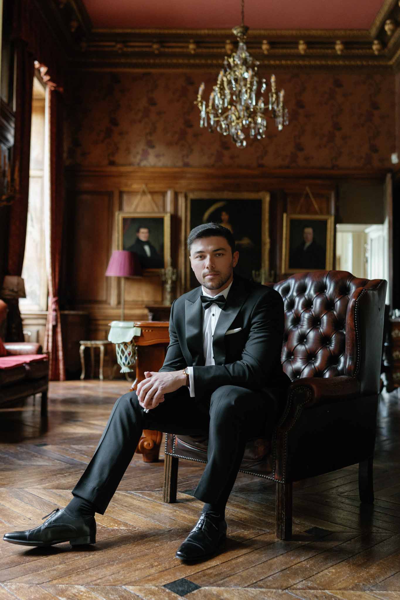 Groom in black tuxedo seated in ornate interior with classical architecture and burgundy decor