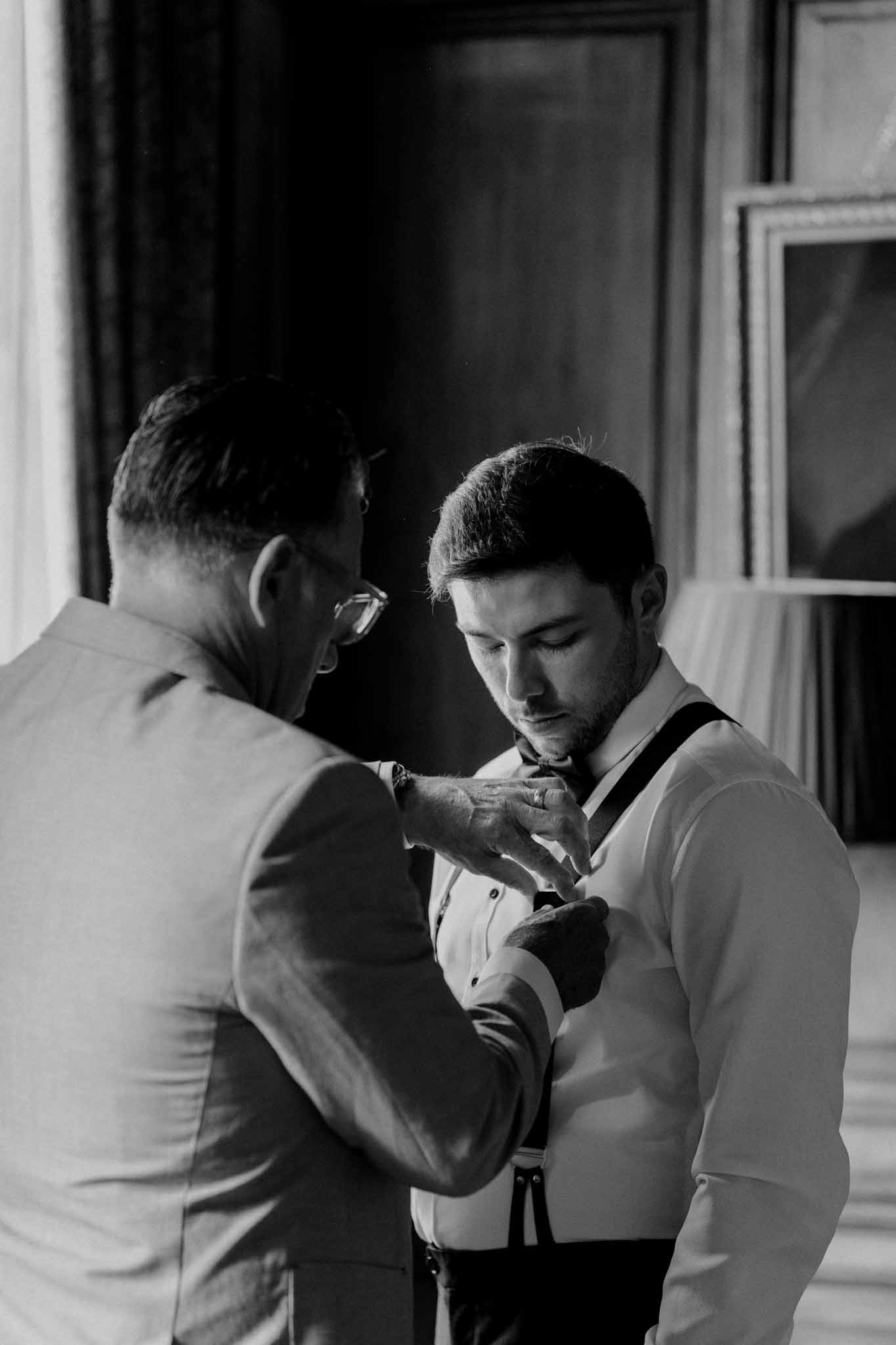 Groom getting ready with groomsman adjusting shirt and vest in interior room