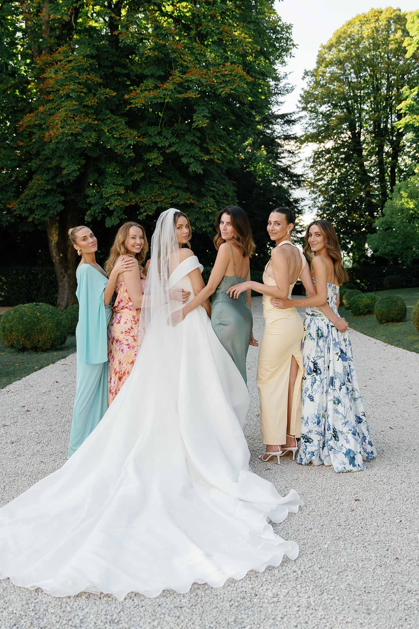 A bridal party portrait taken outdoors on a gravel pathway, likely at a French chateau or estate garden with trimmed box hedges visible in the background. The bride stands at the center wearing an off-the-shoulder white ball gown with a full cathedral-length train and a long sheer veil; the group is posed turning slightly toward the camera. Five bridesmaids or female guests surround her in a mix-and-match approach to dressing: one in a flowing sage-green maxi dress with thin straps, one in a light teal draped one-shoulder style, one in a pink and orange floral printed gown, one in a pale yellow slit dress with a halter neckline, and one in a blue and white floral printed skirt with a strapless top. The styling is modern with a garden-party feel, and the wide portrait shot captures the full length of the bride's train spreading across the gravel path.