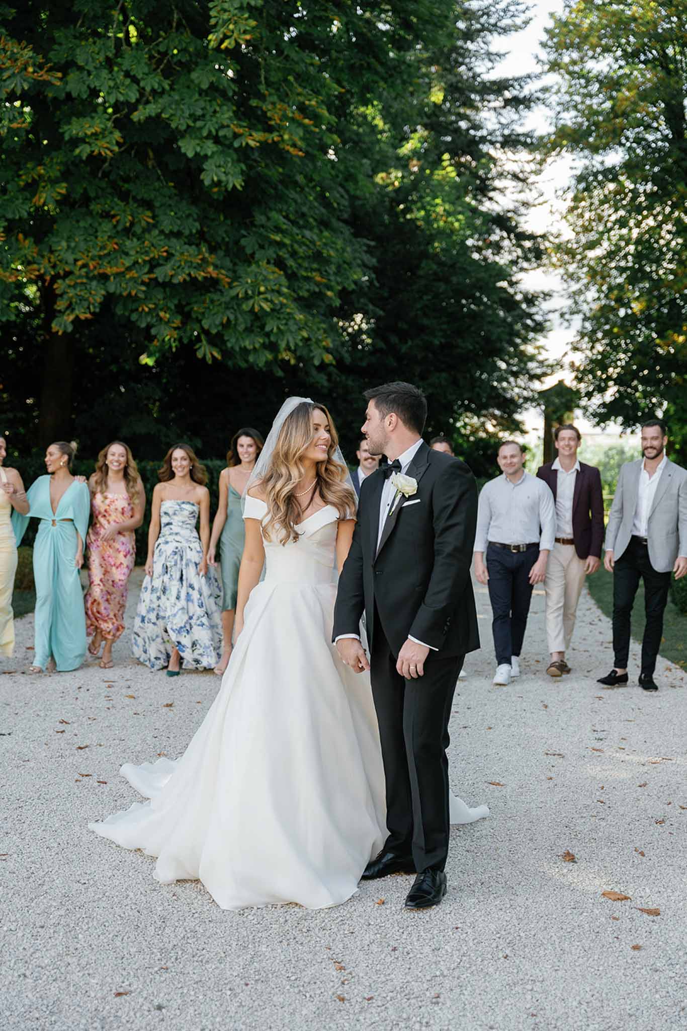 The bride and groom walk hand-in-hand along a gravel path, turning to look at each other, with a group of approximately ten guests following behind them in a candid outdoor portrait. The bride wears an off-the-shoulder ivory ball gown with a full skirt and a sheer veil, with loose wavy hair, while the groom is dressed in a black tuxedo with a bow tie and a cream rose boutonniere. Guests behind them are dressed in a mix of colorful and neutral outfits, including a blue-and-white floral maxi dress, a sage green gown, a teal wrap dress, and various suits and casual smart attire. The setting is an outdoor gravel driveway lined with large mature trees, suggesting a chateau or estate grounds, shot in a wide portrait-style composition with natural evening light.