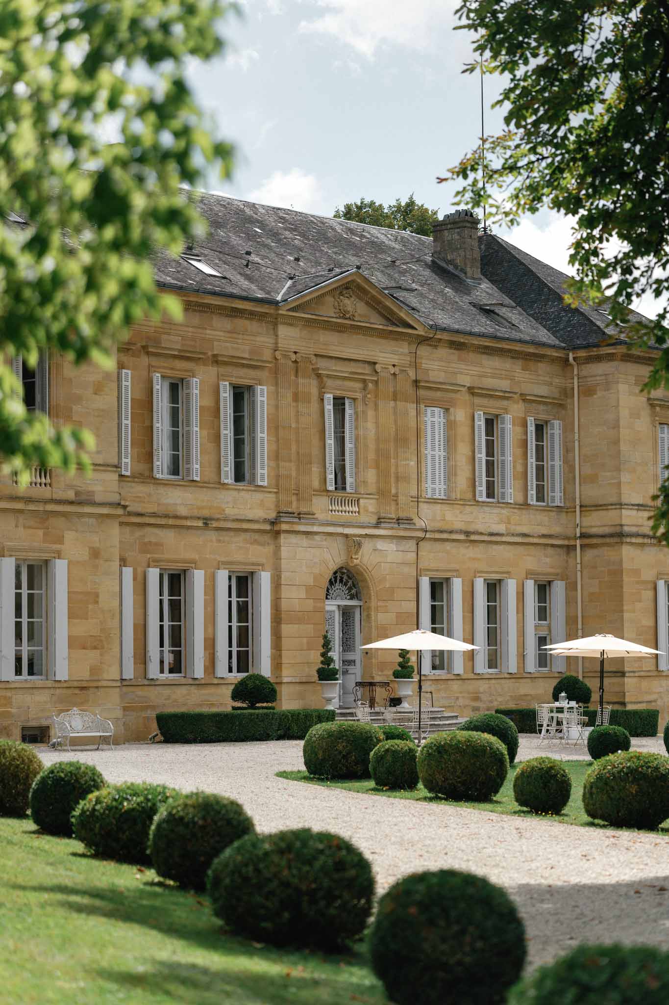 Exterior shot of a French chateau with classical 18th-century architecture, featuring warm golden limestone facades, white shuttered windows, a slate mansard roof, and a decorative pediment above the central entrance with a fanlight door surround. The forecourt features a gravel driveway lined with neatly clipped spherical boxwood topiaries and formal hedging, with a white wrought-iron bench visible to the left. Two cream market umbrellas shade white bistro-style outdoor furniture arranged near the entrance, suggesting a cocktail hour setup. The image is framed by out-of-focus tree foliage in the foreground, shot in portrait orientation as a wide establishing shot. Potential venue feature image.