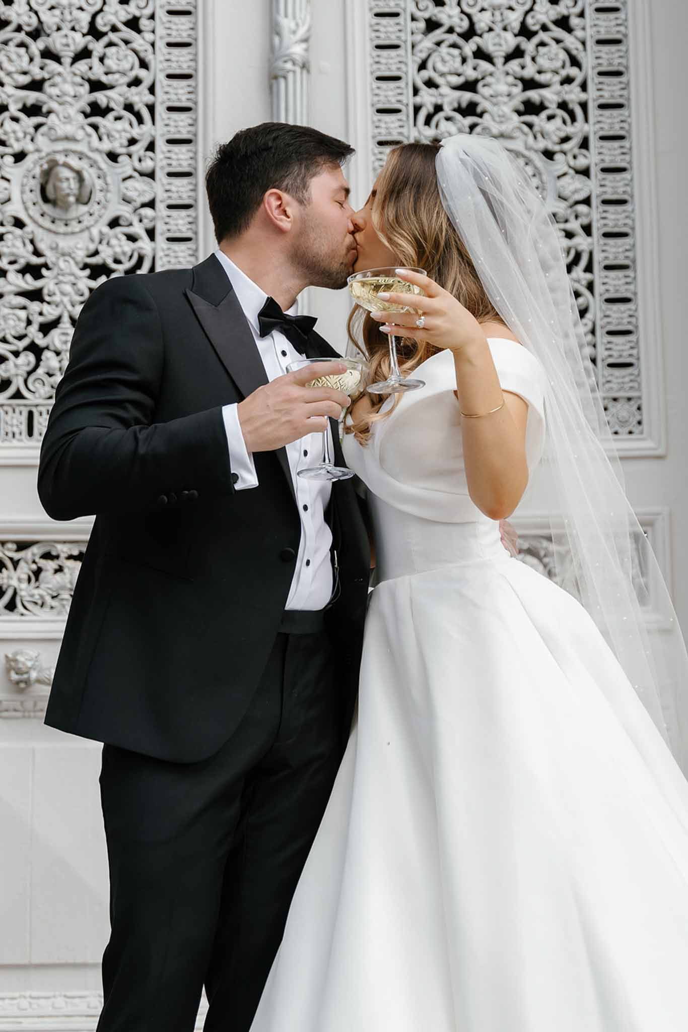 A couple portrait showing the bride and groom kissing while each holding a coupe glass of champagne, photographed indoors against an ornate white carved plasterwork panel with floral and medallion relief detailing. The groom wears a black tuxedo with a black bow tie and white dress shirt with stud buttons, while the bride wears an off-the-shoulder white satin ballgown with a full skirt and a cathedral-length veil with scattered pearl or crystal embellishments. The bride's left hand displays a diamond engagement ring and she wears a delicate gold bracelet. The image is a mid-length portrait shot with a classic, formal styling aesthetic.