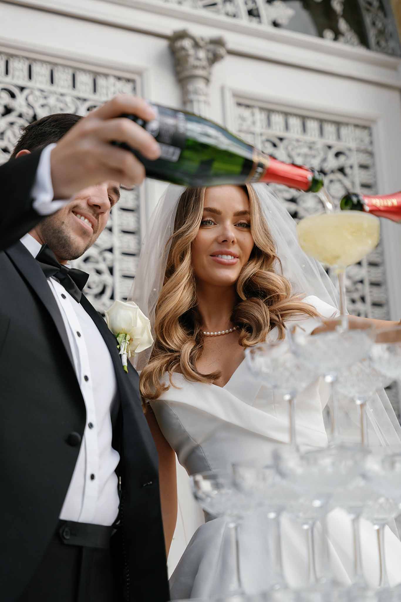 A bride and groom pour champagne into a coupe glass tower during what appears to be a cocktail hour or reception moment outdoors in front of an ornate white building facade with decorative ironwork detailing. The groom wears a black tuxedo with a black bow tie and a white rose boutonniere, while the bride wears an off-the-shoulder white gown with a cathedral-length veil, a pearl necklace, and loose wavy hair. The champagne bottle has a distinctive red foil neck, and the coupe tower contains multiple stacked glasses beginning to fill with sparkling wine. The shot is taken from a low angle looking up, creating a close portrait-style composition that frames both faces alongside the champagne tower in the foreground.
