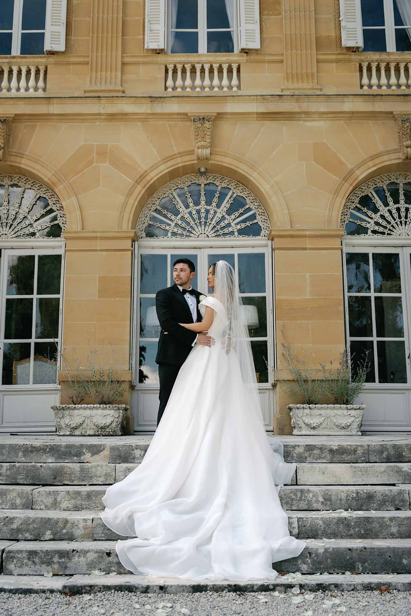 A couple portrait taken outdoors on the stone steps of a French chateau, with the bride and groom posed in an embrace facing each other. The groom wears a black tuxedo with a black bow tie, while the bride wears an off-the-shoulder white ballgown with a voluminous skirt and long cathedral-length train that fans across the steps, paired with a fingertip-length veil. The chateau facade features warm golden limestone, ornate arched doorways with decorative ironwork fan details, white shuttered windows, and carved stone planters flanking the entrance. The composition is a full-length portrait shot from a low angle, emphasizing the dramatic sweep of the gown and the classical architecture behind the couple. The overall styling aesthetic is classic and formal. Potential venue feature image.
