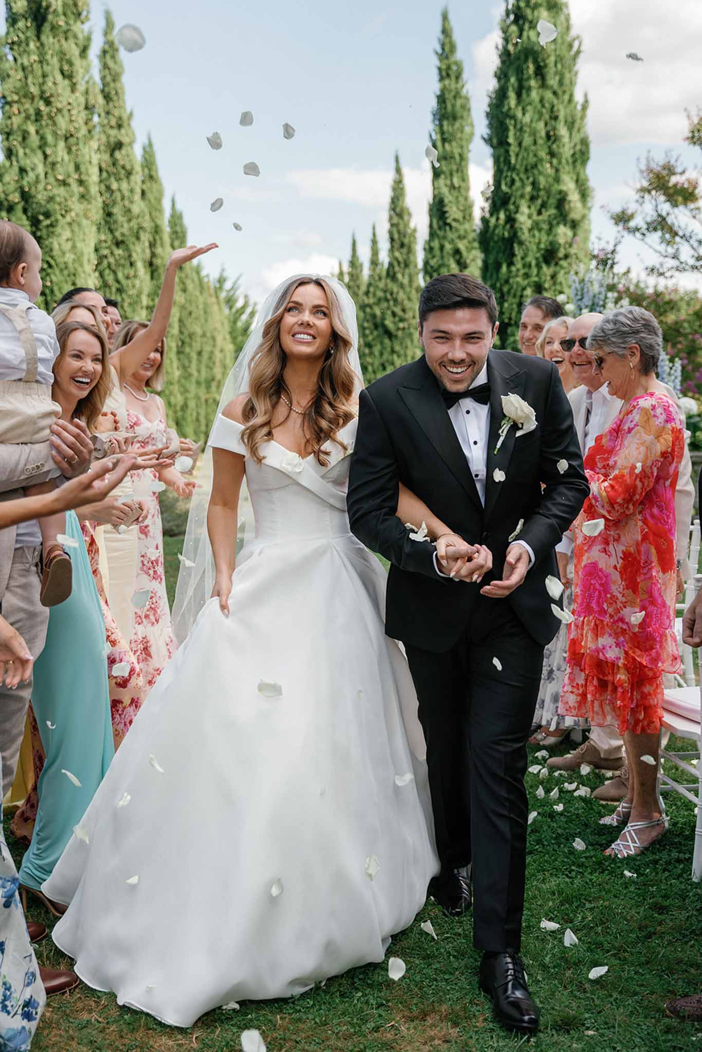 The couple walks back down the aisle during an outdoor ceremony recessional, surrounded by approximately 20 guests lining both sides who are tossing white rose petals into the air. The setting is a garden with tall Italian cypress trees forming a backdrop. The bride wears an off-the-shoulder white ball gown with a voluminous skirt and a long veil, with wavy loose hair and a delicate necklace; the groom wears a black tuxedo with a bow tie and a white rose boutonniere. Guests are dressed in colorful summer attire including bold floral print dresses in pink and red, a mint green dress, and neutral linen suits. The composition is a mid-shot taken at ground level, capturing the couple at center frame with guests blurred on either side.