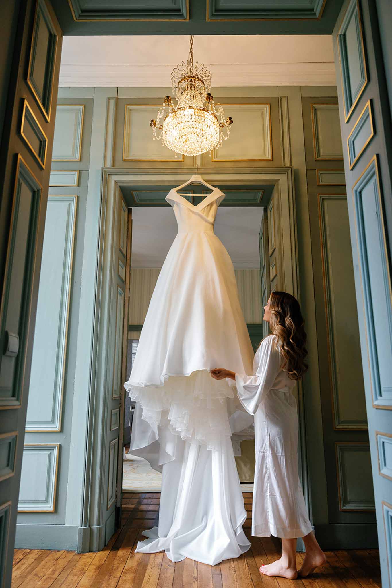 Bride in white robe admiring wedding dress in classical hallway with sage doors and gold chandelier