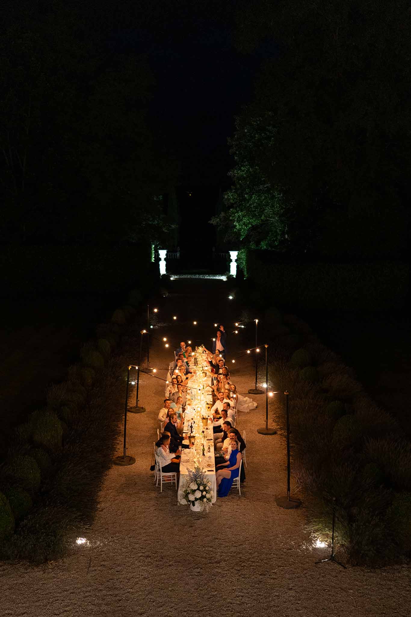 An aerial wide shot of an outdoor wedding reception dinner set at night in what appears to be a formal garden pathway of a French château or estate. Approximately 30–40 guests are seated along a single long rectangular table lined with white linens, candlelight, and a white floral centerpiece with blue accents visible at the near end. The table runs along a gravel allée flanked by clipped topiary hedges, with a stone balustrade structure illuminated in the background. Warm bistro-style string lights on freestanding poles run alongside the table on both sides, providing the primary ambient lighting alongside the candles on the table. The overall styling is classic and formal, with guests dressed in dark suits and formal attire including at least one cobalt blue dress visible near the foreground.