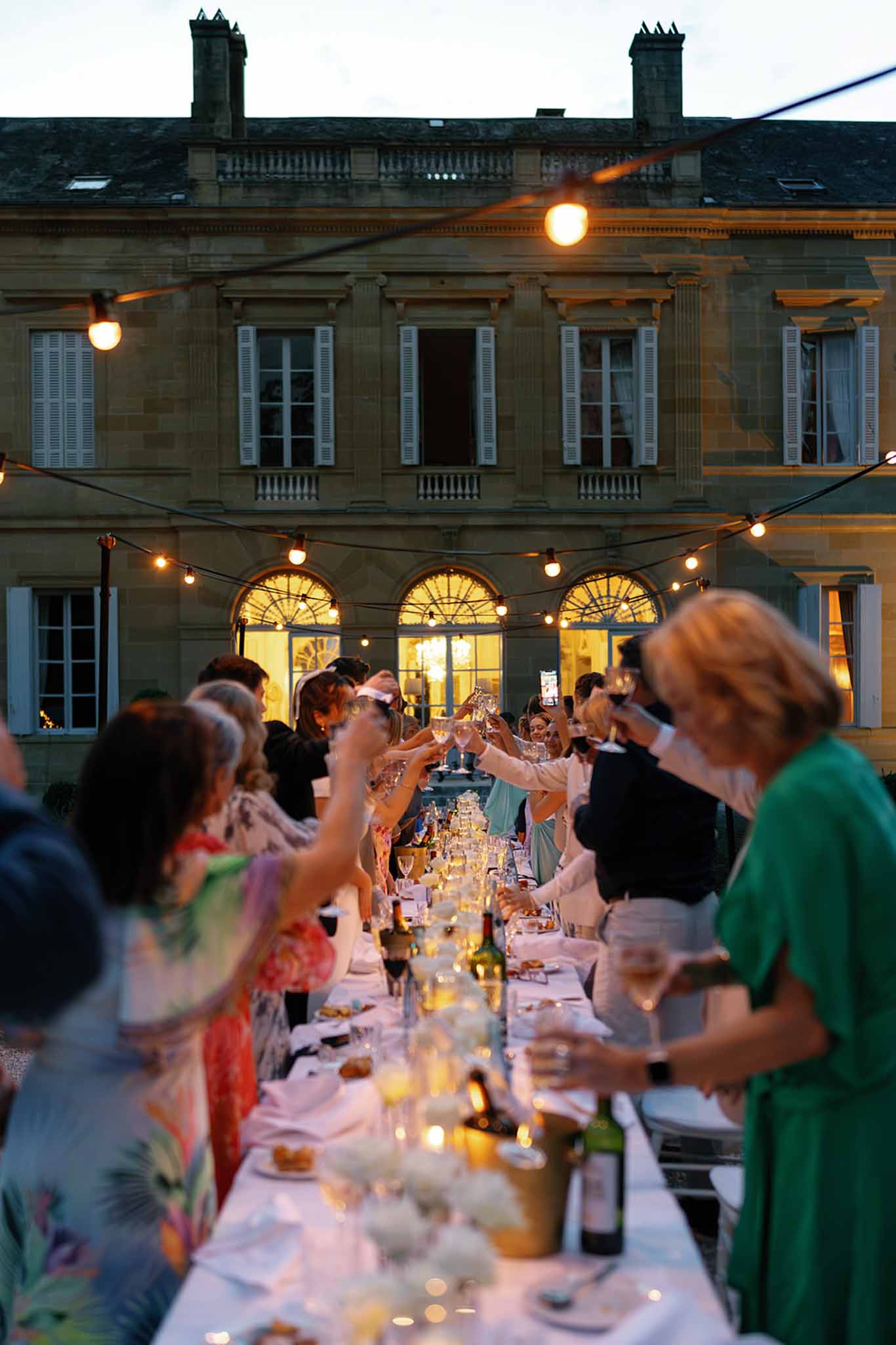 An outdoor wedding reception dinner is taking place in the courtyard of a French chateau at dusk, with approximately 20–30 guests seated and standing along a single long banquet table covered in a white linen tablecloth. Guests are raising their glasses in a toast, with wine bottles, candles in votives, and what appear to be low white floral arrangements running down the center of the table. The guests are dressed in colorful cocktail attire, including a floral-print dress and a kelly green dress visible in the foreground. Overhead Edison bulb string lights are strung across the courtyard, casting warm amber light, while the chateau's arched ground-floor windows glow golden from interior lighting. The shot is taken from one end of the table looking toward the chateau facade, creating a strong perspective line — a wide, slightly shallow-depth-of-field shot that keeps the foreground guests soft and the chateau in focus in the background. Potential venue feature image.