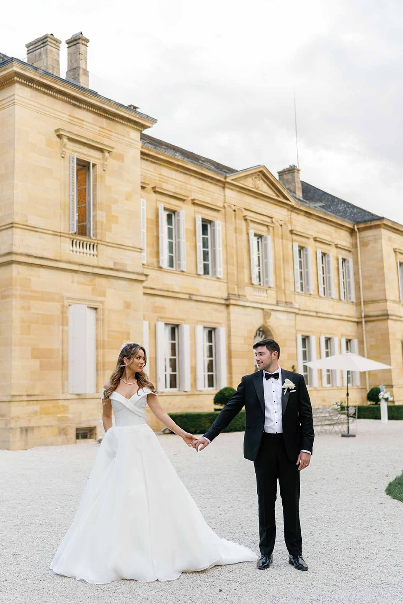 A couple portrait taken outdoors on the gravel forecourt of a French château, a large honey-colored limestone manor house with white shuttered windows, a slate roof, and clipped topiary hedges visible in the background. The bride wears an off-the-shoulder white ball gown with a full skirt and train, while the groom wears a black tuxedo with a white dress shirt, black bow tie, and a white rose boutonnière. They are holding hands and looking at each other, with the bride slightly turned away from camera. The shot is a full-length portrait with the château facade filling the background. Potential venue feature image.