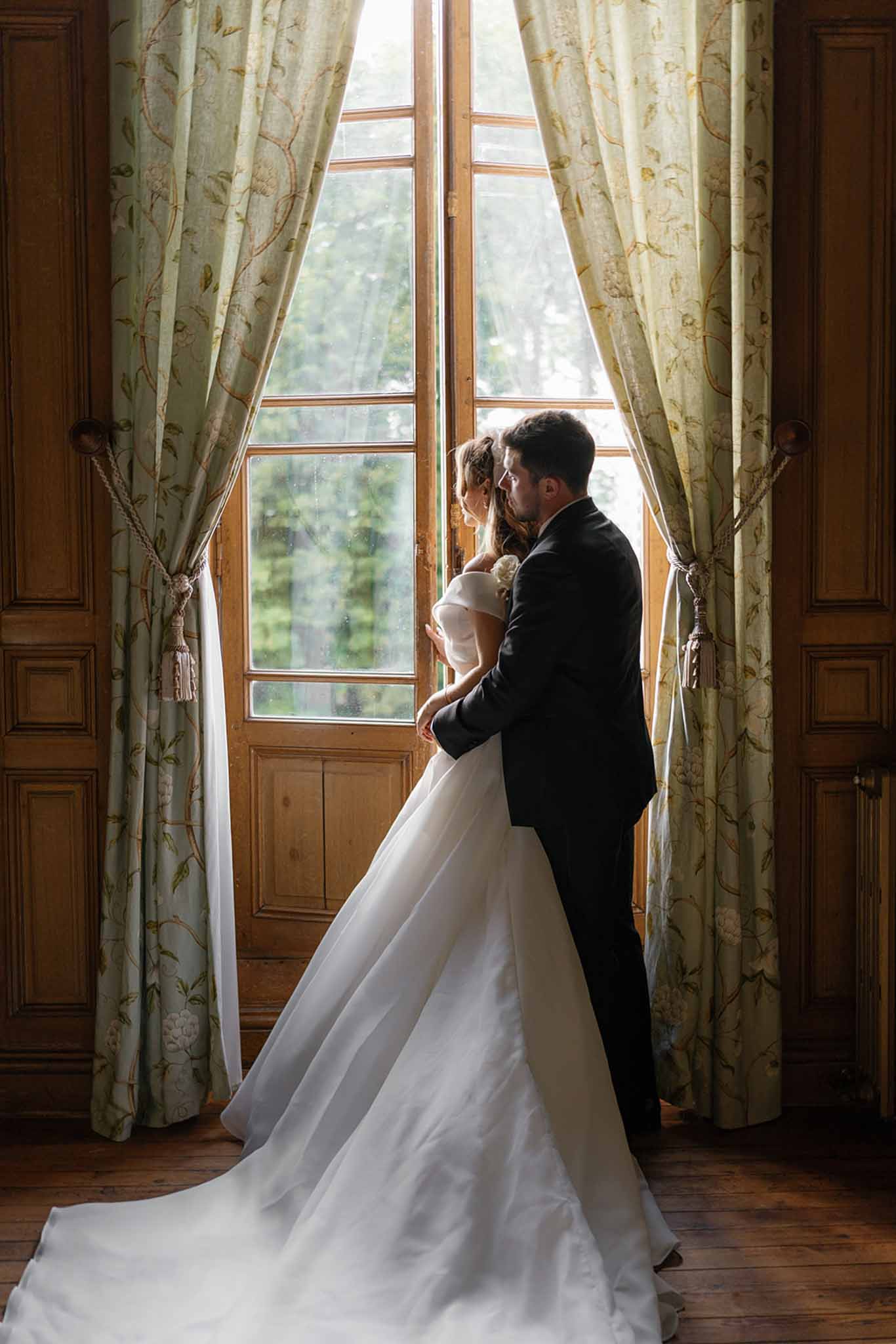 A couple portrait taken indoors, likely inside a French chateau, with the bride and groom standing together in front of tall French doors framed by sage green floral-patterned curtains with rope-and-tassel tiebacks. The groom, wearing a dark navy suit, stands behind the bride with his arms around her waist, their faces close together. The bride wears an off-the-shoulder white ballgown with a long train that pools across the dark hardwood floor, and her hair is loosely pinned up. Natural backlight streams through the glass-paned doors, creating a warm contrast against the wood-panelled walls of the room. The composition is a full-length portrait shot from a slight distance, centering the couple symmetrically between the curtains.