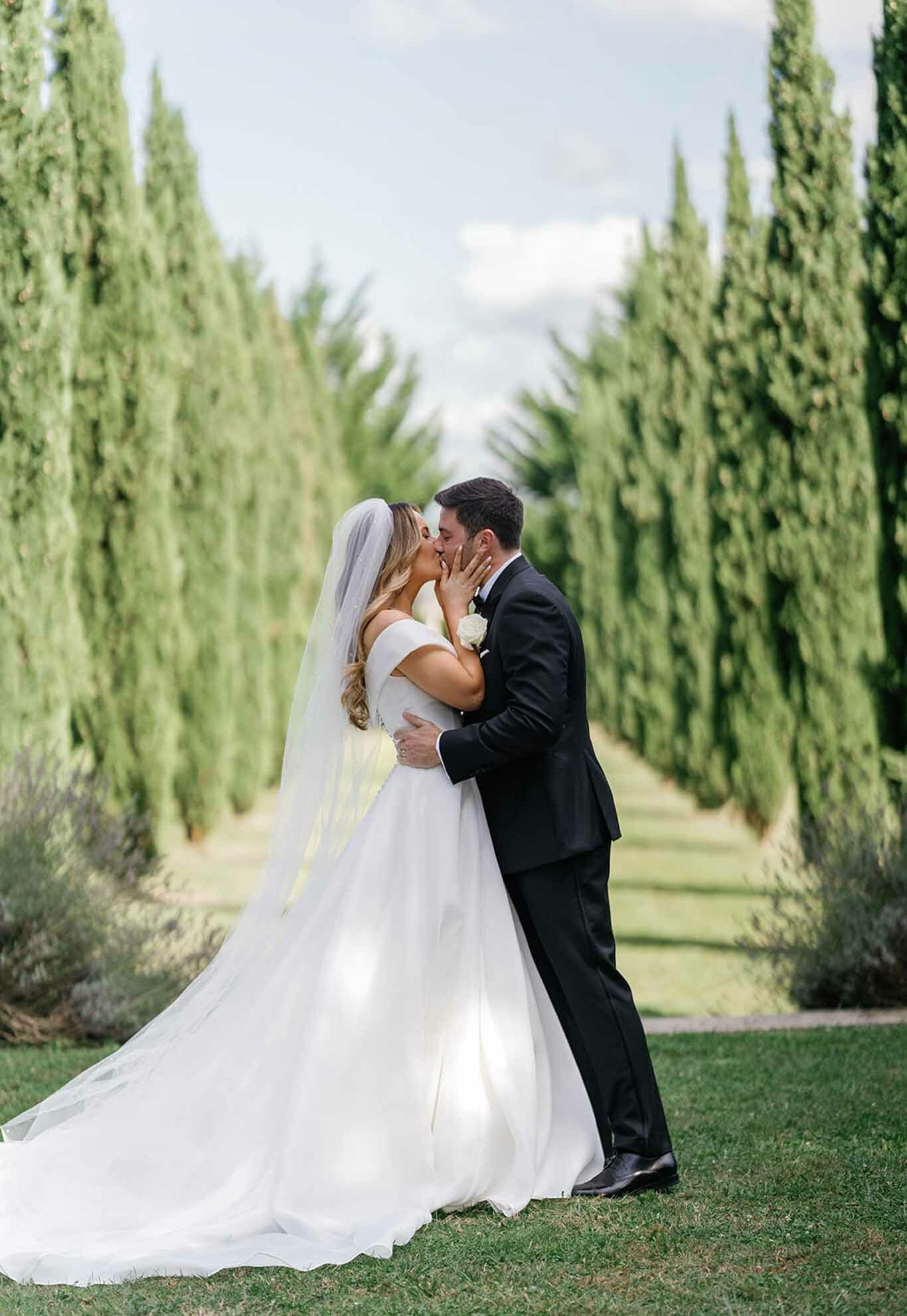 A couple portrait shot outdoors in a formal garden allee lined with tall, narrow Italian cypress trees on both sides, creating a strong perspective leading into the background. The bride and groom are kissing at the center of the frame; the bride wears an off-the-shoulder white ball gown with a full skirt and cathedral-length veil, with loose blonde waves, while the groom is dressed in a classic black tuxedo with bow tie and a white rose boutonniere. The composition is a full-length portrait with the cypress allee providing a symmetrical backdrop, styled in a clean, classic aesthetic with lavender bushes visible at the base of the trees.