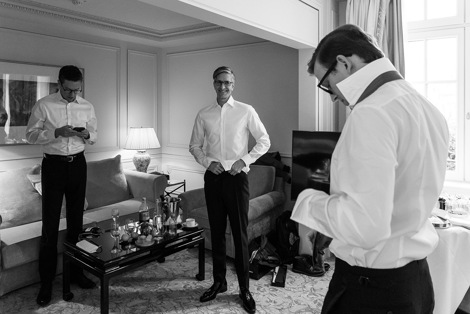 A black-and-white getting-ready scene showing three men in a well-appointed hotel suite, all in the process of dressing. The man in the center, likely the groom, smiles while adjusting his shirt cuffs; the man on the left checks his phone; and the man in the foreground on the right adjusts his shirt sleeve and tie. All three wear white dress shirts and dark trousers. The room features paneled walls, a pleated table lamp, a upholstered sofa with cushions, and a coffee table holding water bottles, champagne in an ice bucket, wine glasses, and a coffee cup. Dress shoes and bags are visible on the floor near the sofa. The image is a wide-angle candid shot with high contrast between the bright shirts and the darker tones of the trousers and furniture.