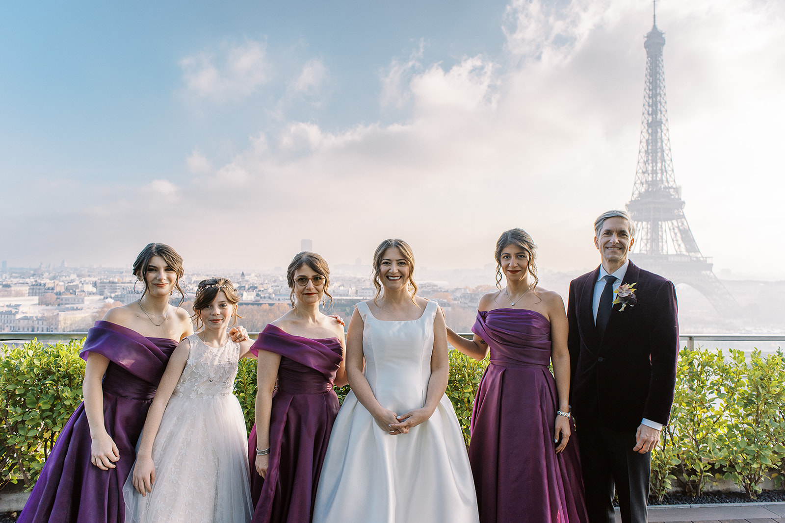 A bridal party group portrait taken on an outdoor rooftop terrace in Paris, with the Eiffel Tower prominently visible in the background along with a wide panoramic view of the city. The bride stands at center in a structured, sleeveless ivory satin ball gown with a square neckline, flanked by three bridesmaids wearing deep purple off-the-shoulder satin gowns with draped wrap-style bodices. A young flower girl in a white floral-embroidered dress stands between the bridesmaids on the left side, and a man in a dark burgundy suit with a navy tie and a small floral boutonniere stands at the far right. The group of six faces the camera and the shot is a wide, medium-length portrait framed to include the full Eiffel Tower rising behind them.