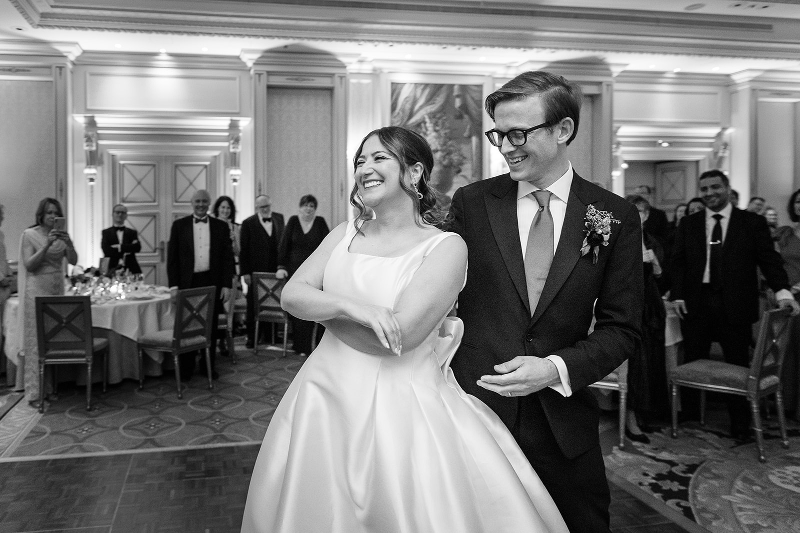 This black-and-white image captures the bride and groom's grand entrance or first dance at an indoor reception held in a formal ballroom with ornate paneled walls, pilasters, and wall sconces providing warm ambient lighting. The bride wears a structured satin ball gown with a square neckline and a large back bow detail, while the groom is dressed in a dark suit with a light-toned tie and a small floral boutonnière. Both are laughing and walking arm in arm, clearly reacting to something in the room, with approximately 15–20 formally dressed guests visible in the background watching and some photographing on phones. The setting features round dining tables with white linens, cross-back chairs, and a patterned carpet, suggesting a classic, formal reception aesthetic. The image is a medium-wide shot with the couple centered in the foreground and guests forming a natural frame behind them.