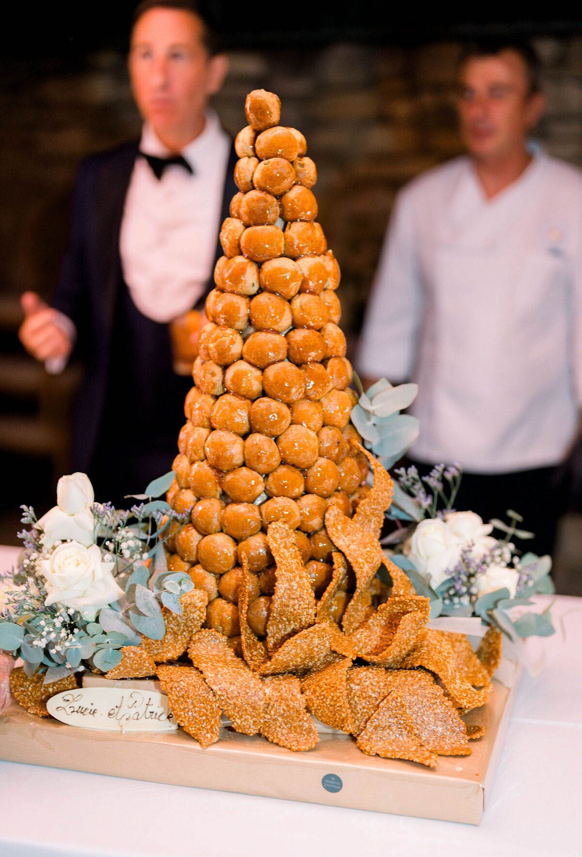 A traditional French croquembouche wedding cake is displayed at a reception, built as a tall cone-shaped tower of caramel-glazed choux pastry profiteroles surrounded by spun caramel and nougatine sugar decorations at the base. The cake sits on a wooden board on a white-clothed table and is flanked on both sides by small floral arrangements of white roses, baby's breath, lavender, and eucalyptus. A small name tag reading 'Lucie et Patrice' is placed at the front of the board. In the soft-focus background, two men are visible — one in a navy tuxedo with a black bow tie, the other in a white chef's jacket — suggesting a cake presentation moment during the reception. The shot is a medium close-up focused on the dessert with shallow depth of field.