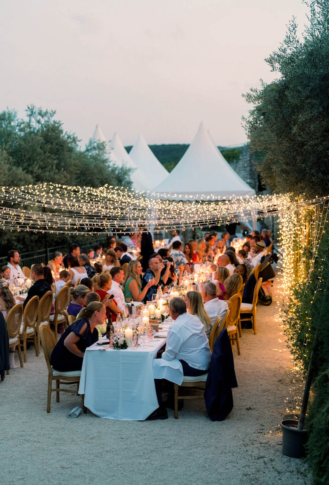 An outdoor wedding reception dinner is underway at dusk, with approximately 60–80 guests seated at long rectangular tables covered in white linen, arranged along a gravel terrace. The tables are lit with clusters of white pillar candles and set with glassware and place settings, with small white floral arrangements visible. Guests are dressed in a mix of smart-casual attire including white shirts, navy dresses, and colorful summer outfits. Overhead, dense canopies of warm white fairy lights are strung across the entire dining area and draped over trimmed hedges and potted topiaries lining the right side, creating a warm ambient glow against the dusky evening light. Rattan-back gold chairs are used throughout. In the background, three white peaked marquee tent tops are visible alongside a stone building, suggesting a French countryside or Provence-style venue. The shot is a wide, slightly elevated portrait-orientation image capturing the full length of the reception setup.