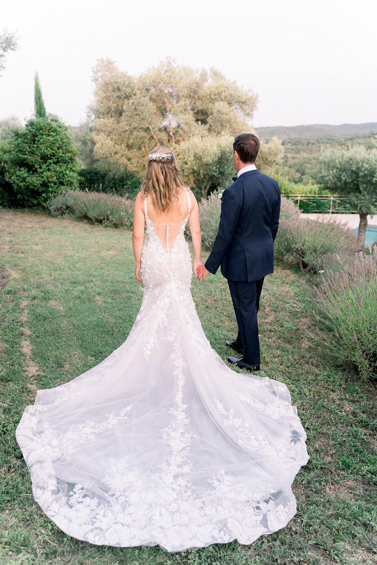 A couple portrait taken outdoors in a garden setting, shot from behind as the bride and groom walk hand in hand away from the camera. The bride wears a fitted mermaid-silhouette gown in ivory with an illusion low-cut back featuring floral lace appliqués and a very long cathedral train that spreads across the ground; her hair is worn down in loose waves with a floral and crystal hair piece. The groom wears a navy suit with a white boutonniere. Lavender borders line the path beside them, with a pool visible in the background and rolling hills in the distance. The composition is a full-length portrait that prominently showcases the back of the bridal gown and its train.