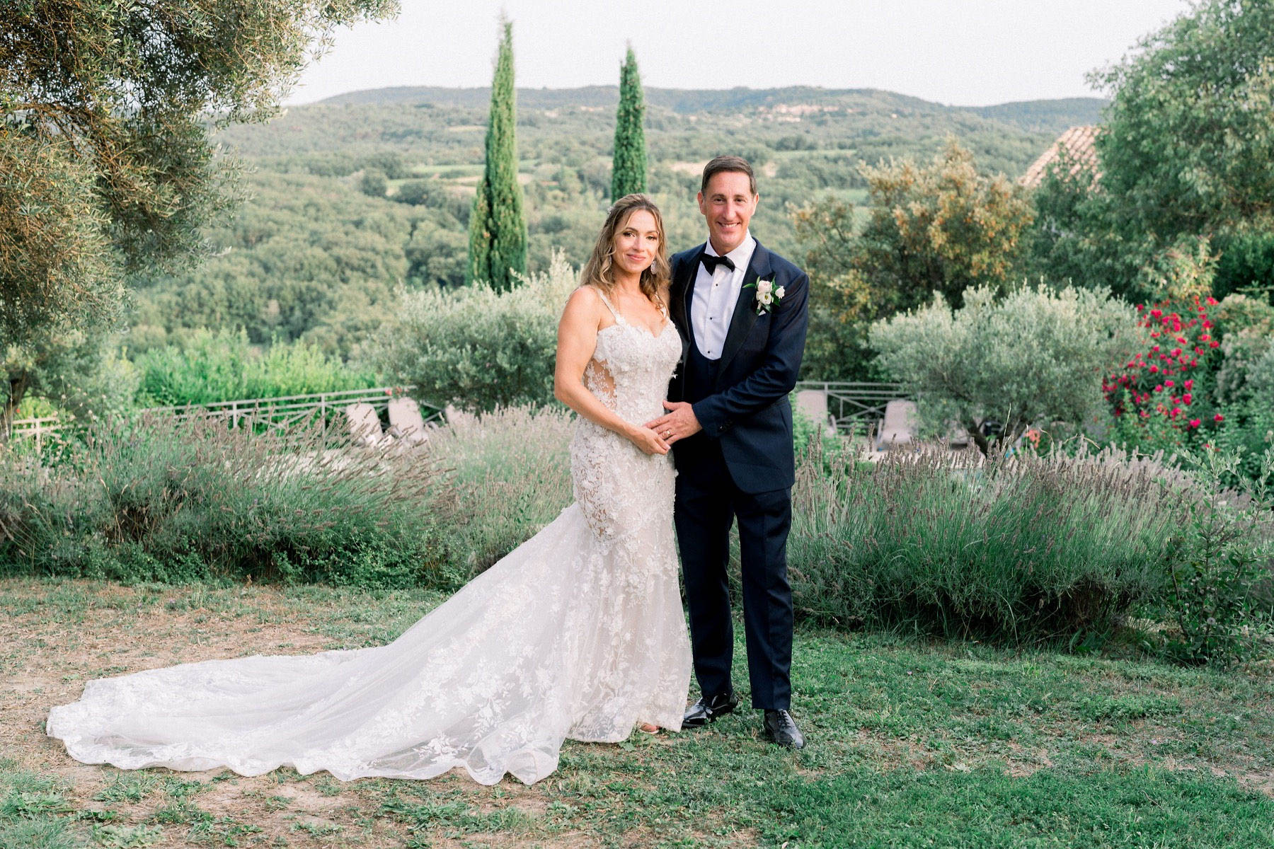 A couple portrait taken outdoors in a French countryside garden setting, with rolling hills and cypress trees visible in the background. The bride wears a fitted ivory lace mermaid gown with spaghetti straps, a deep V-neckline, floral appliqué detailing, and a long cathedral train; the groom wears a navy tuxedo with a black bow tie and a white rose boutonniere. The couple stands close together facing the camera, with rows of lavender and climbing red roses visible in the background garden. This is a mid-length portrait shot with a soft, natural light typical of late afternoon.
