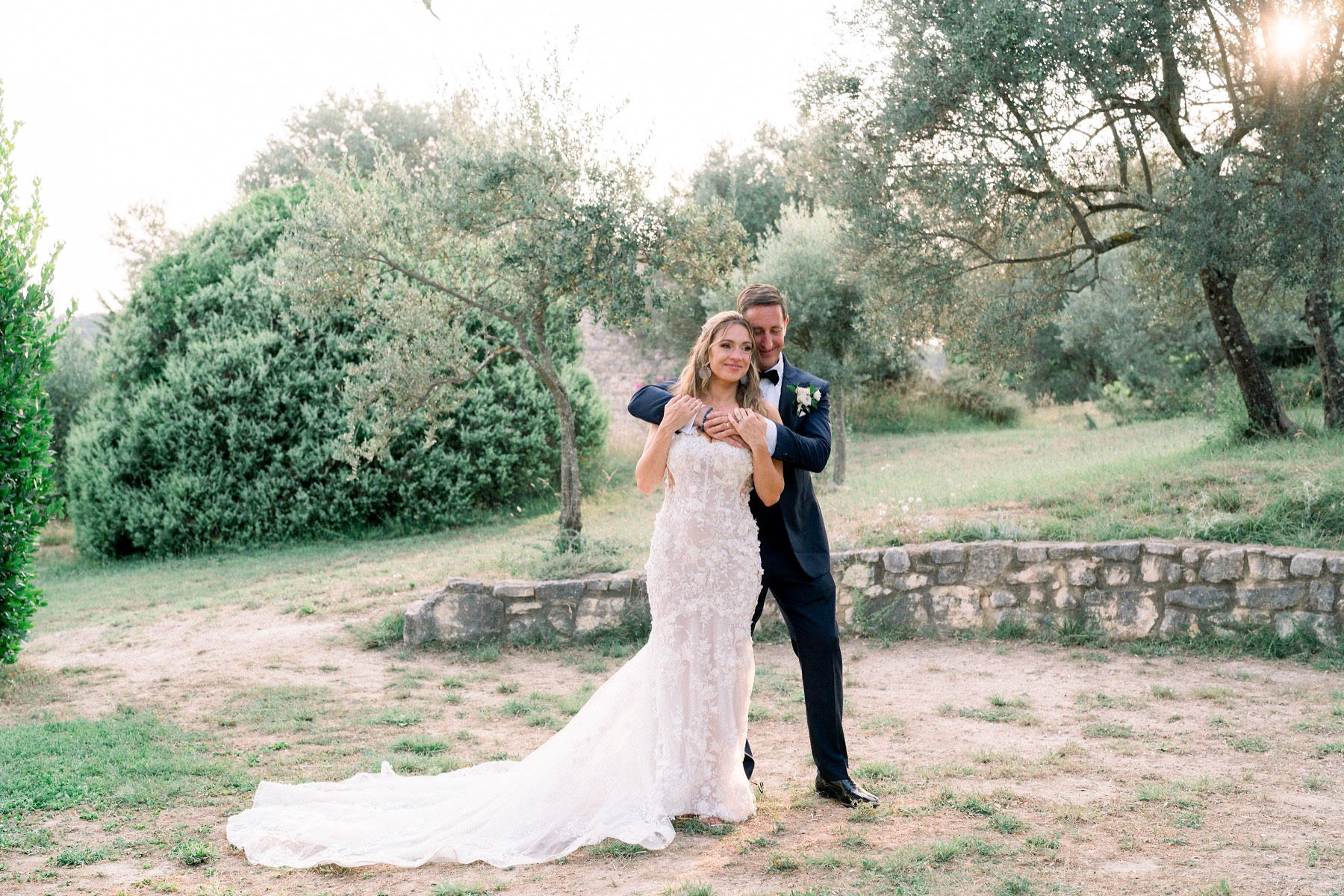 A couple portrait taken outdoors during golden hour, set among olive trees with a low stone wall visible in the background. The groom stands behind the bride with his arms wrapped around her, both smiling toward the camera. The bride wears a fitted, floor-length ivory lace gown with floral appliqué detailing and a trailing train, while the groom is dressed in a navy tuxedo with a black bow tie and a white boutonniere. The warm backlit light creates a soft glow across the scene, giving the image a bright, airy quality. The composition is a mid-length couple portrait shot.