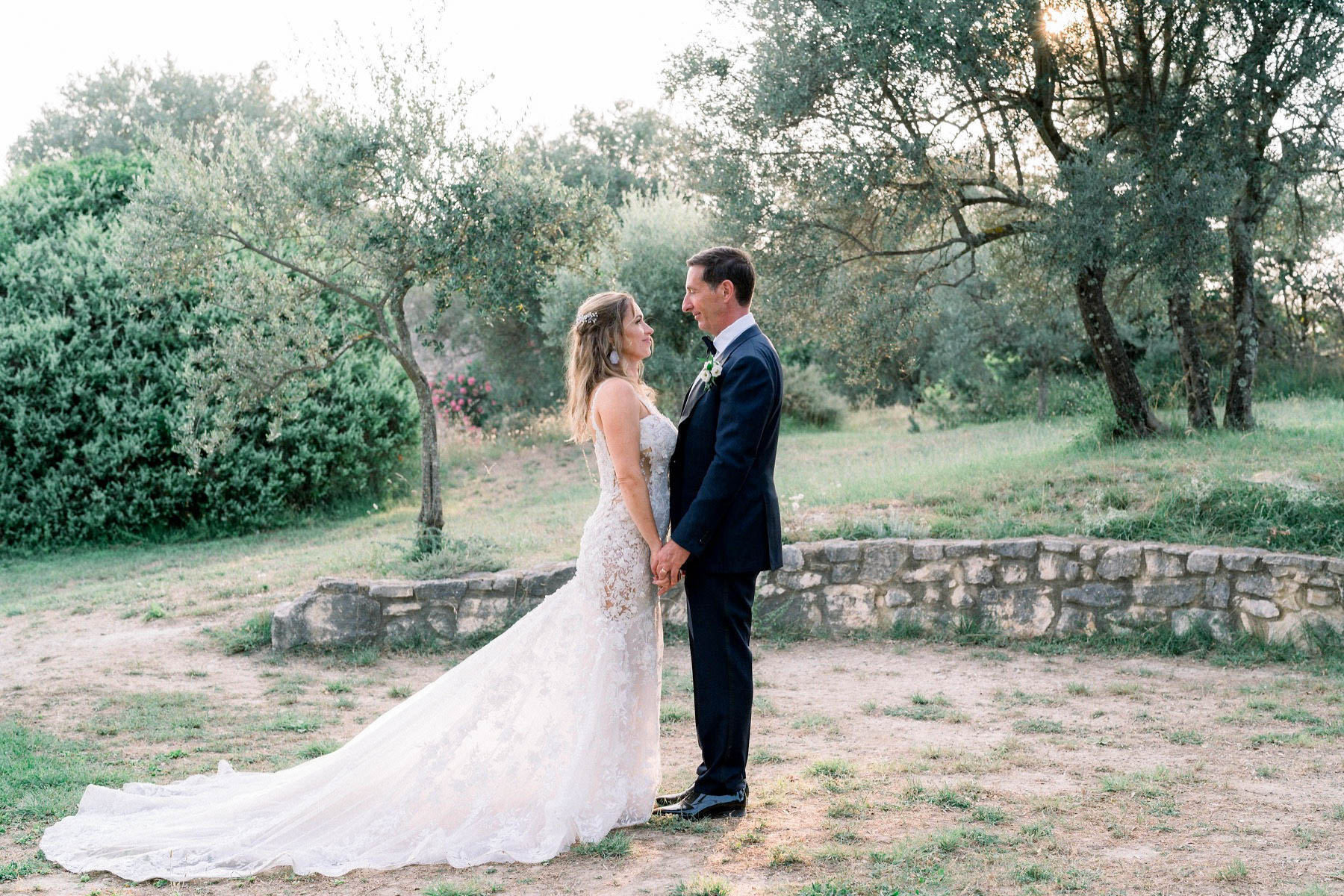 A couple portrait taken outdoors among olive trees, with the bride and groom facing each other and holding hands. The bride wears a fitted ivory lace gown with a long cathedral train and a delicate floral hair accessory, while the groom wears a navy suit with a white boutonniere and black bow tie. The setting appears to be the grounds of a French countryside property, with a low dry-stone wall visible in the background. The shot is a medium full-length portrait taken in warm late-afternoon light.