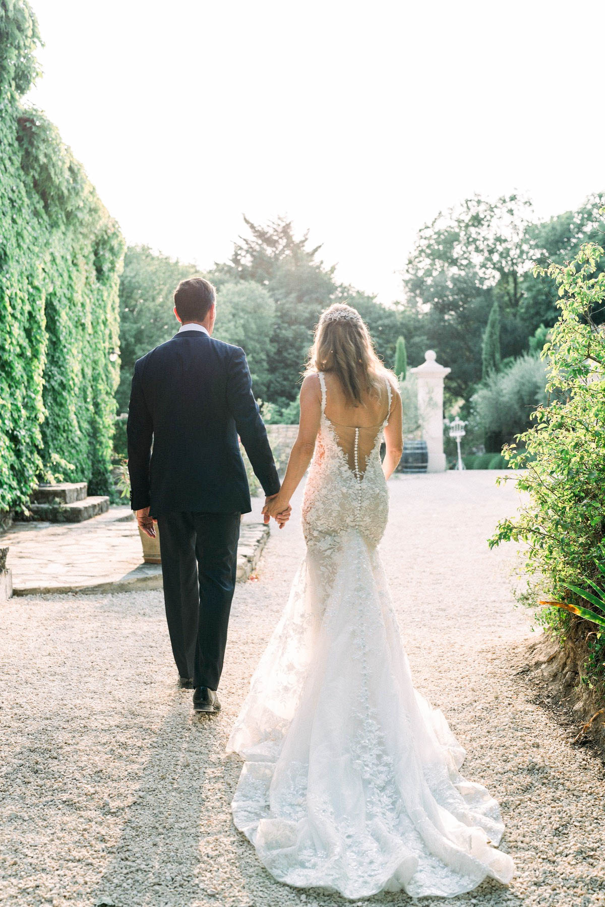 A couple portrait taken from behind as the bride and groom walk hand-in-hand along a gravel path in an outdoor garden setting. The groom wears a navy suit, while the bride wears a fitted ivory lace gown with a deep open back detailed with a row of button closures down the spine and a full trailing train. Her hair is worn down with a pearl or crystal hair accessory at the crown. A decorative stone gate pillar is visible in the background, suggesting a formal garden or chateau grounds. The image is shot in a wide portrait composition with warm golden-hour backlight, giving the scene a soft, airy quality.