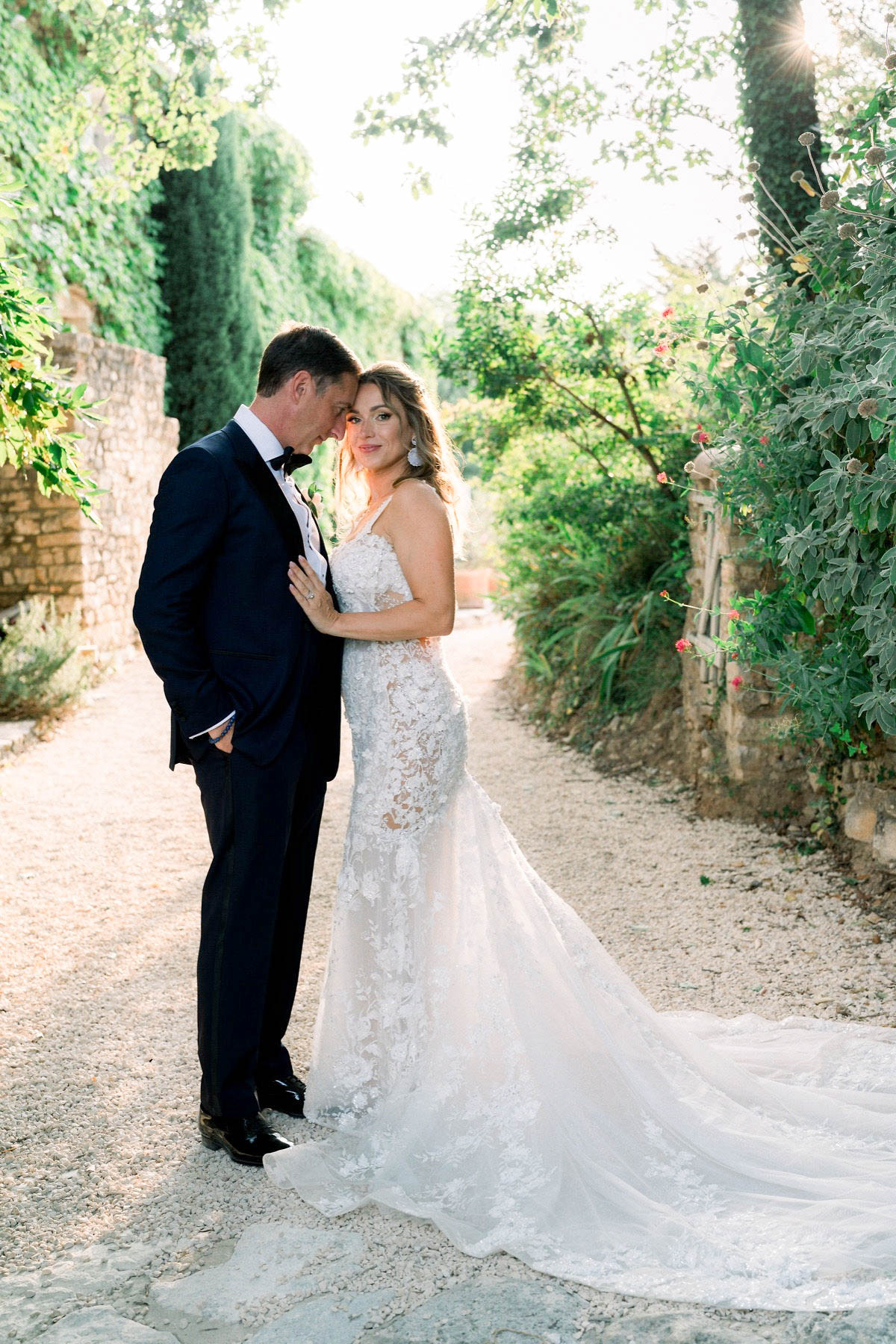 A couple portrait taken outdoors on a gravel garden path flanked by stone walls covered in climbing plants. The bride wears a fitted ivory lace mermaid-style gown with floral appliqué, spaghetti straps, and a long trailing train; she accessorizes with drop earrings and rests her hand on the groom's chest while looking toward the camera. The groom wears a navy tuxedo with a black bow tie and a small boutonniere, and leans his forehead toward the bride's. The shot is a full-length portrait composed along the garden path, lit by warm golden backlight suggesting late afternoon sun.