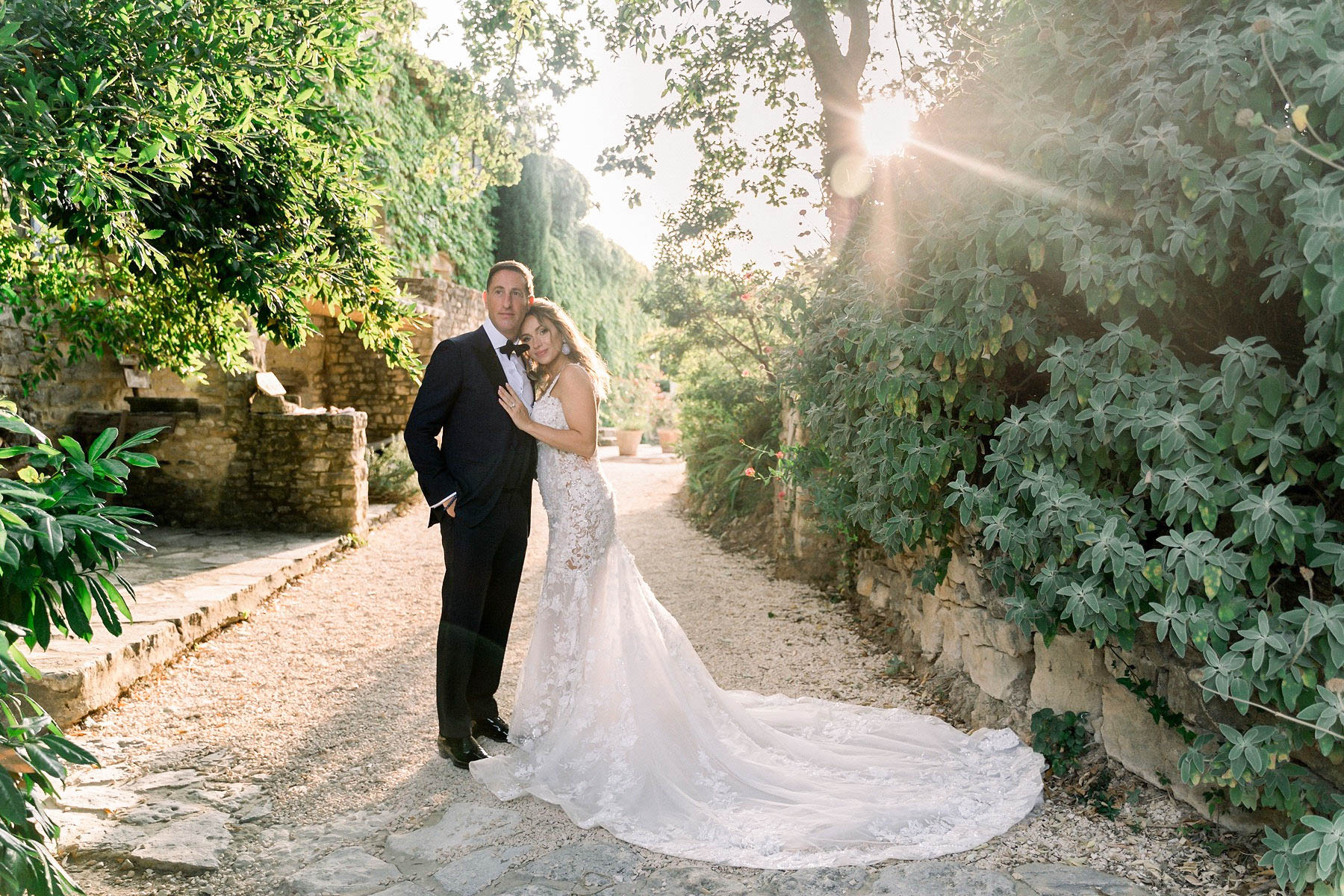 A couple portrait taken outdoors along a gravel pathway flanked by ivy-covered stone walls at golden hour, with a visible sun flare breaking through the tree canopy on the right. The groom wears a classic black tuxedo with a black bow tie, while the bride wears a fitted ivory lace mermaid gown with intricate floral appliqué and a long cathedral train spread across the gravel path. The bride leans her head against the groom's cheek with one hand resting on his chest, and both face the camera. The setting has a classic French countryside feel with aged stone architecture visible in the background. The shot is a medium-wide portrait composition.