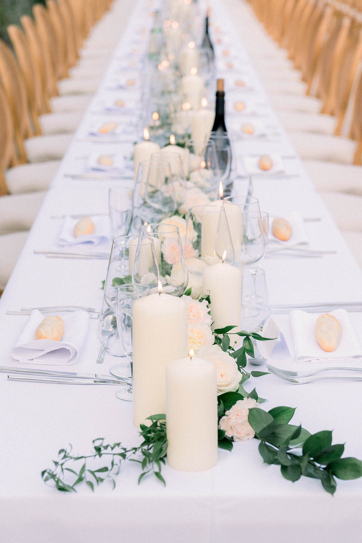 A detail shot of a long rectangular reception table set for a wedding dinner, photographed from a low angle to emphasize the length of the table receding into the background. The white linen tablecloth is dressed with a central garland runner of green eucalyptus and ruscus foliage interspersed with ivory pillar candles of varying heights, blush carnations, and cream garden roses. Each place setting includes a white folded napkin topped with a small oval peach-toned item, clear stemmed wine glasses, and polished silver cutlery. Natural wood cross-back chairs line both sides of the table. The overall decor palette is white, ivory, blush, and green, consistent with a classic minimalist styling approach.