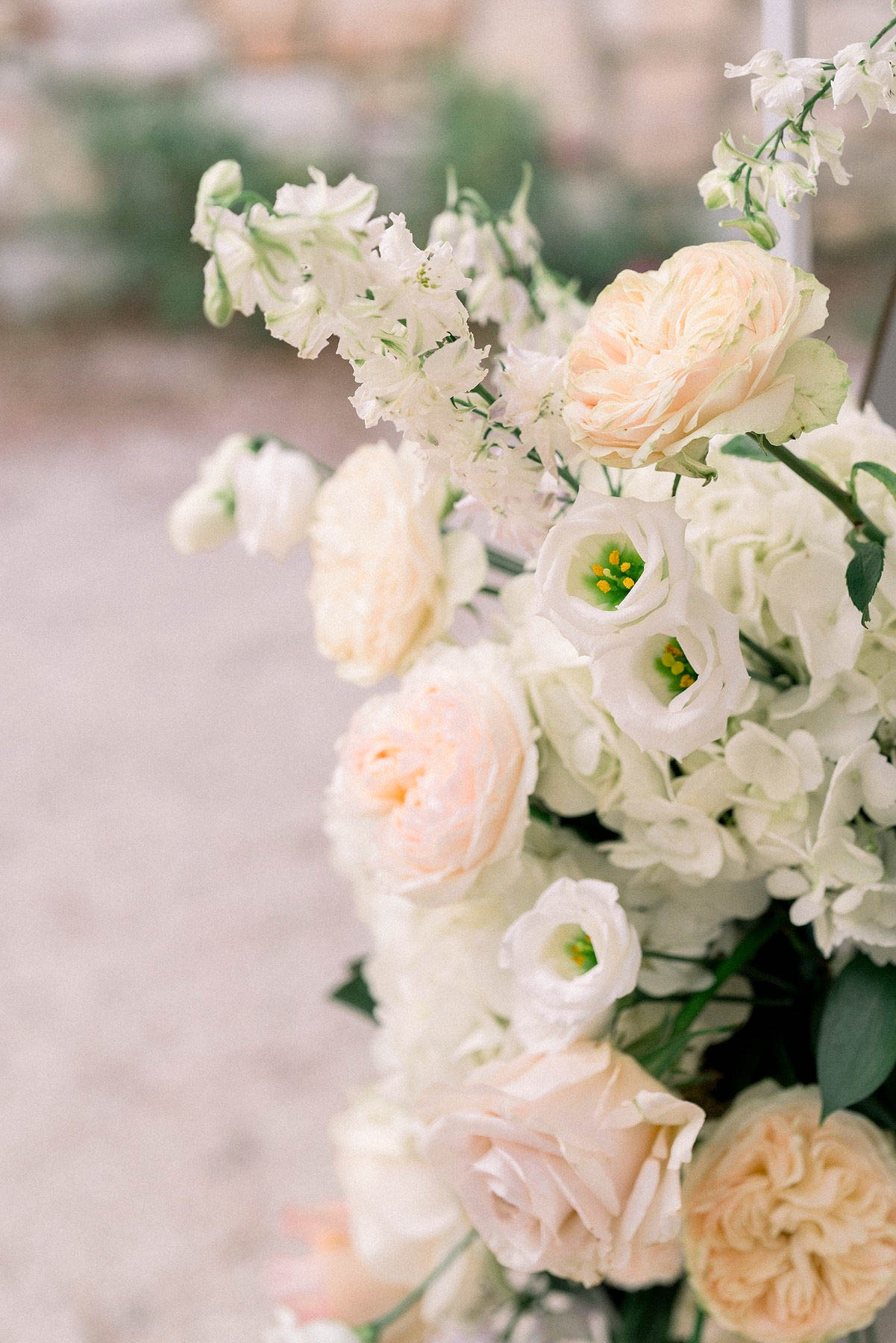 Close-up detail shot of a floral arrangement featuring blush and peach garden roses, white lisianthus with visible yellow stamens, white hydrangea, and white delphinium spires, accented with green foliage. The color palette is soft and pale, combining ivory, blush, peach, and cream tones in a classic romantic style. The arrangement appears to be an outdoor ceremony floral installation, likely an aisle marker or altar piece, with a gravel surface visible in the blurred background. The image uses a shallow depth of field, keeping the foreground blooms sharp while the background falls out of focus.