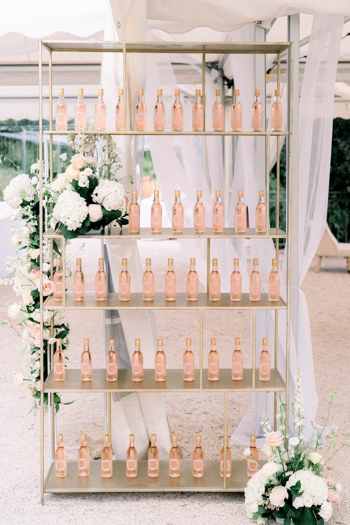 A wedding favor or escort card display featuring a tall five-shelf gold metal open bookcase holding approximately 40 miniature rosé wine bottles with custom white-printed labels and gold caps. The display is set under a white tent or marquee with sheer white fabric draping in the background. The left side of the shelving unit is decorated with a lush floral arrangement of white hydrangeas, white stock flowers, blush garden roses, and greenery, with a smaller coordinating floral cluster at the bottom right corner. The overall decor palette is blush, white, and gold, consistent with a classic, romantic wedding aesthetic. This is a medium wide shot capturing the full display.