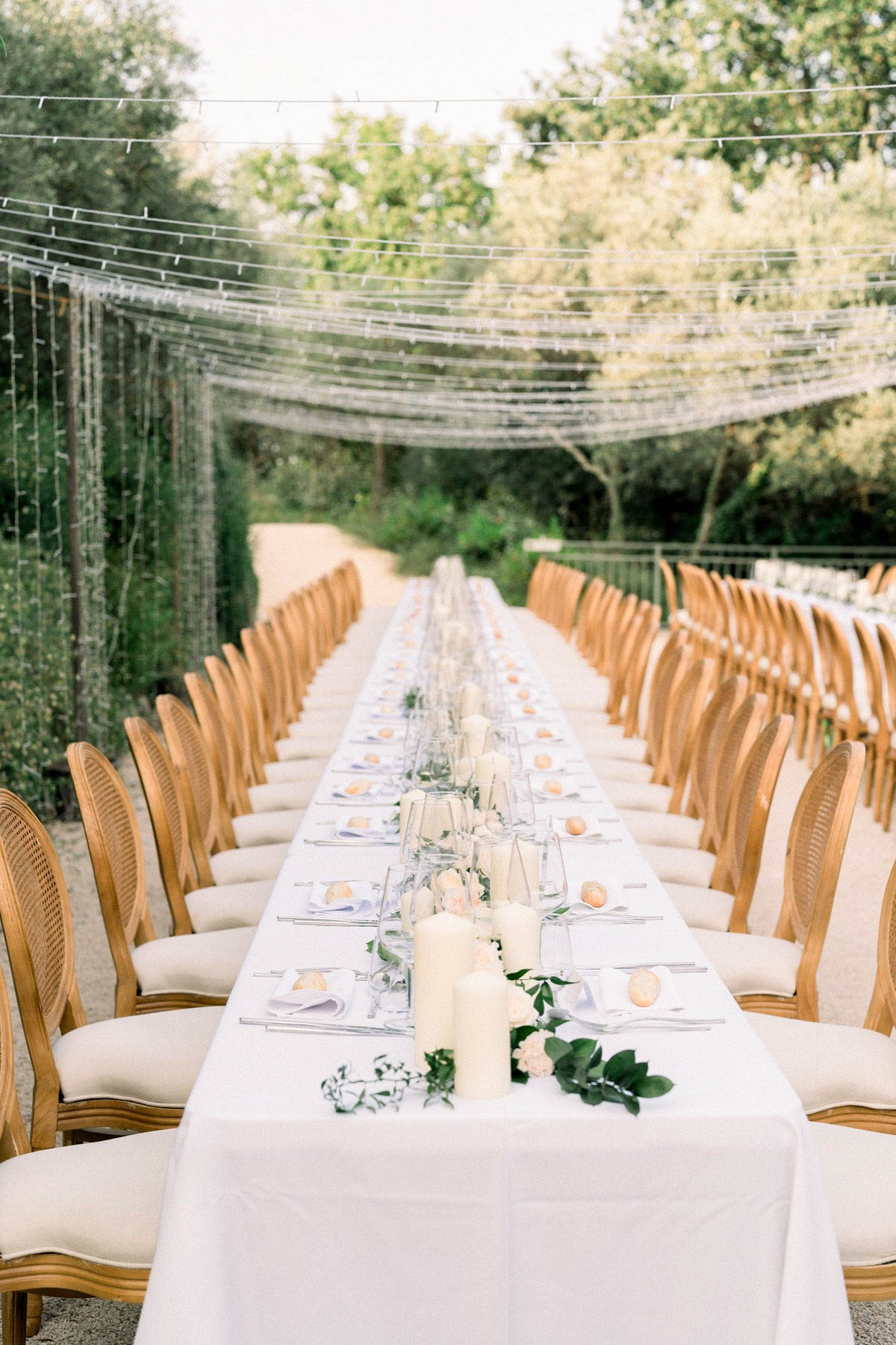 An outdoor wedding reception table setup photographed in a wide perspective shot down the full length of a long feasting table seating approximately 40 guests. The table is dressed with a white linen tablecloth, lavender-tinted folded napkins, clear glassware including champagne flutes, silver cutlery, and small round orange citrus fruits used as decorative place card holders. The centerpiece runs the length of the table with clusters of ivory pillar candles in varying heights, small blush roses, and trailing greenery including eucalyptus and broad-leaf foliage. Natural wood Louis XVI-style cane-back chairs with cream upholstered seats line both sides of the table. Overhead, a dense canopy of fairy lights is strung across a pergola-style structure, creating a grid pattern above the dining area. The overall decor palette is white, ivory, and soft blush with natural wood tones, consistent with a classic outdoor garden reception style.