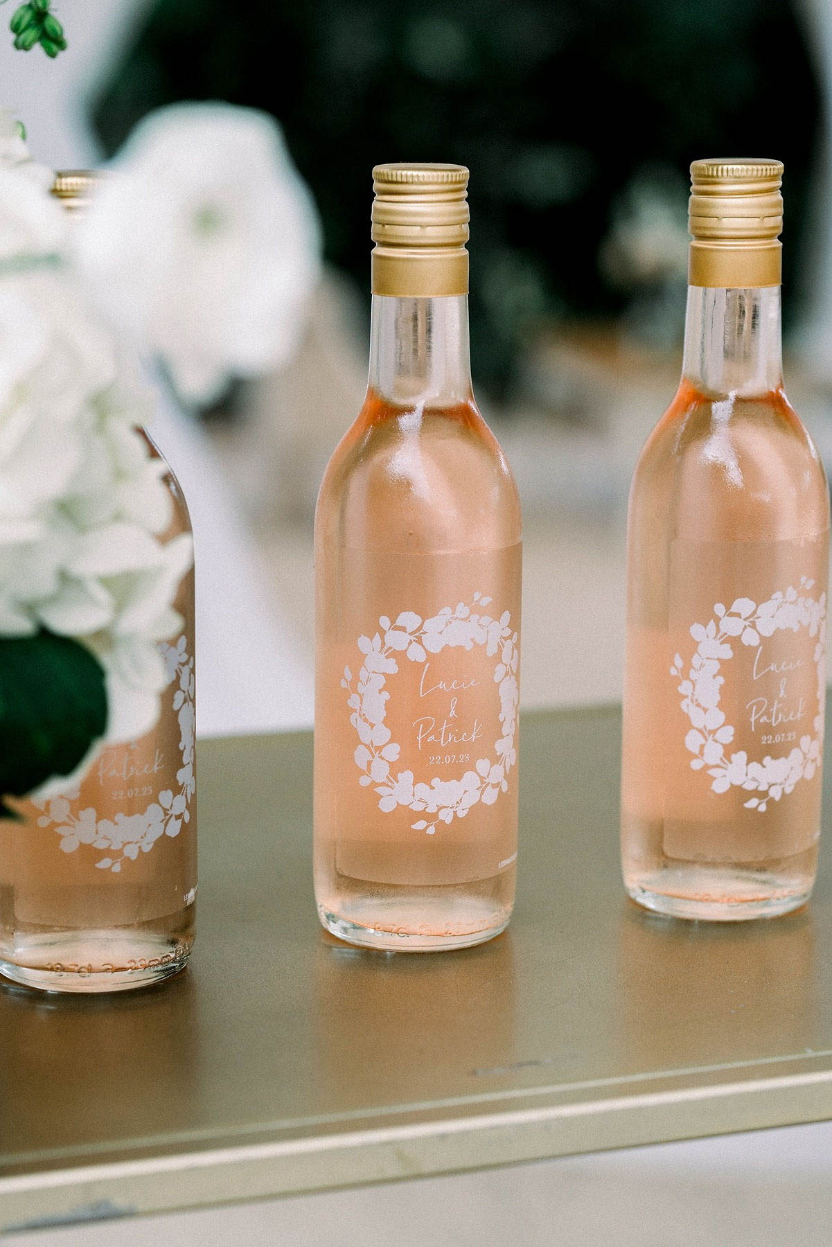 Close-up detail shot of three miniature rosé wine bottles used as wedding favors, displayed on a gold surface. Each bottle contains pale pink rosé wine, is sealed with a gold screw cap, and features a custom white label printed with a floral wreath motif surrounding the text 'Lucie & Patrick 22.07.23'. A partial arrangement of white hydrangeas is visible in the soft-focus background to the left. The overall decor palette combines blush pink, white, and gold, reflecting a classic and polished styling approach.