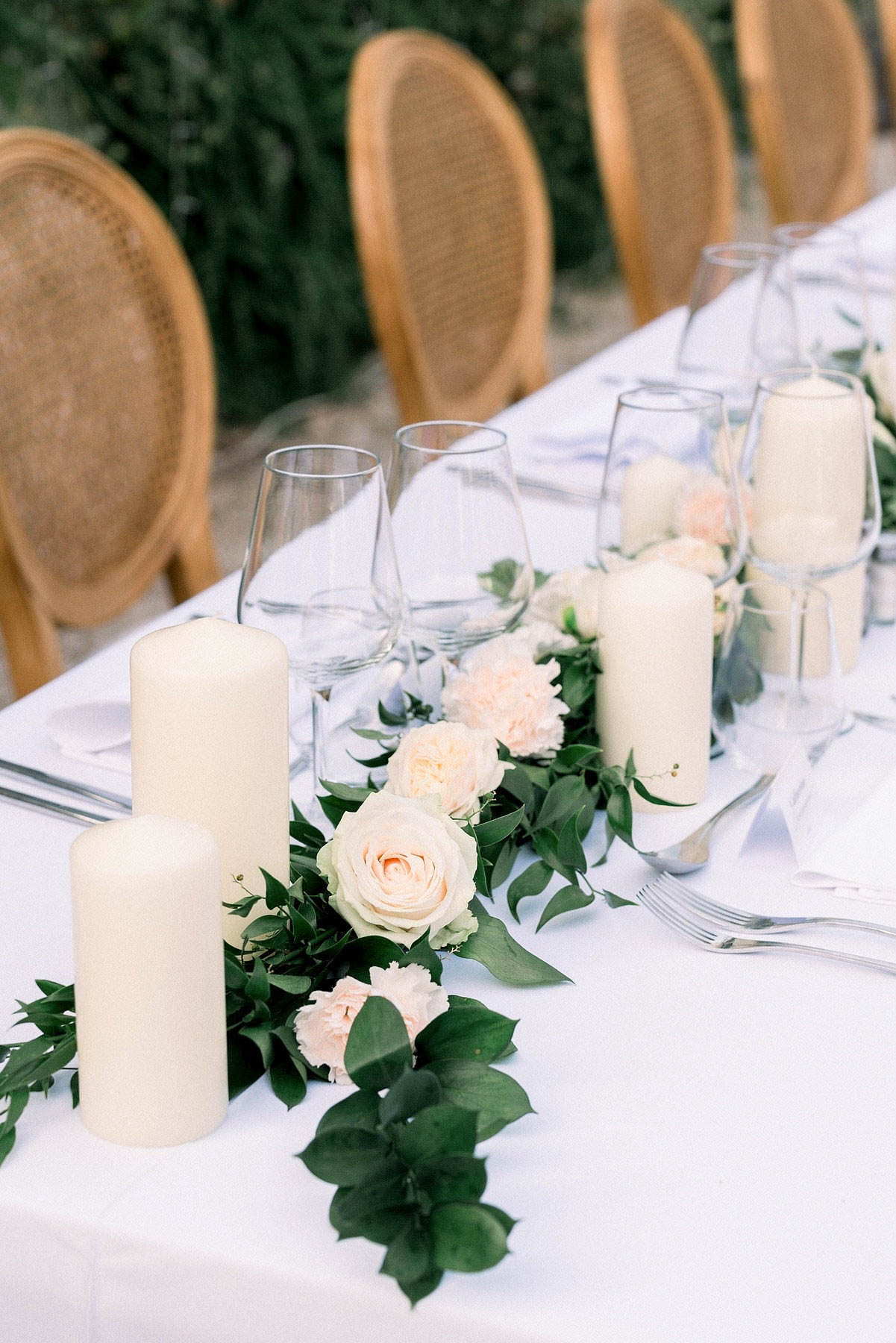Close-up detail shot of an outdoor wedding reception table set with a white linen tablecloth and a garland centerpiece running along the center. The garland features fresh greenery, blush pink carnations, and cream garden roses interspersed with tall ivory pillar candles of varying heights placed in clear glass cylinder holders. Place settings include silver cutlery, white folded napkins, and clear stemless wine glasses. Natural wood rattan-back chairs are visible in the background, contributing to a classic French garden aesthetic with a soft, neutral color palette of white, cream, and blush.