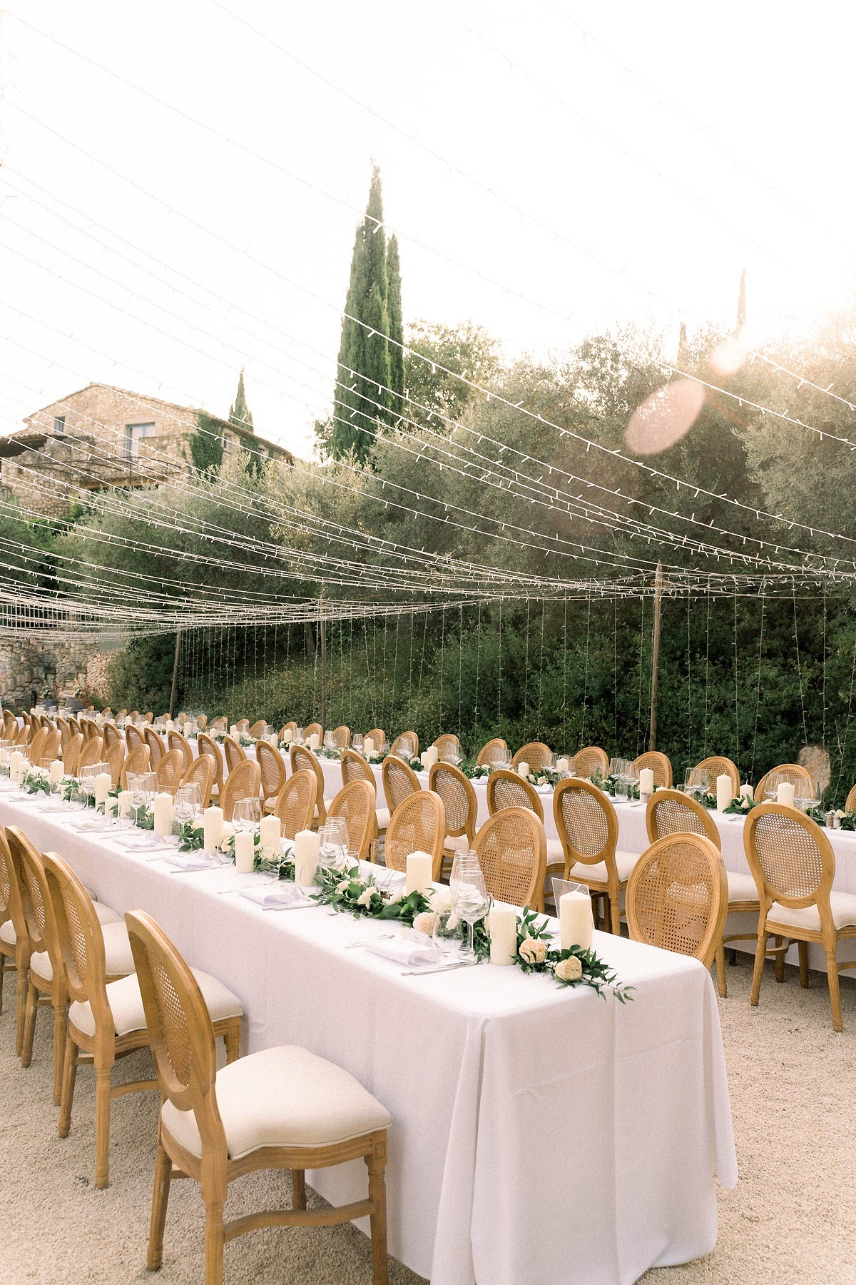 An outdoor wedding reception setup at what appears to be a Provençal stone estate, photographed as a wide shot with visible lens flare from strong backlight. Two long rectangular banquet tables are dressed in white linen tablecloths and lined with natural wood Louis XVI-style cane-back chairs with cream upholstered seats. The table runners consist of trailing greenery with ivory pillar candles of varying heights and small white and pale blush roses interspersed throughout. Place settings include wine glasses, folded grey linen napkins, and flatware. Overhead, an extensive canopy of fairy lights is strung across the dining area, cascading downward in curtain-like vertical strands. The setting is a gravel courtyard with a classic French country aesthetic. Potential venue feature image.