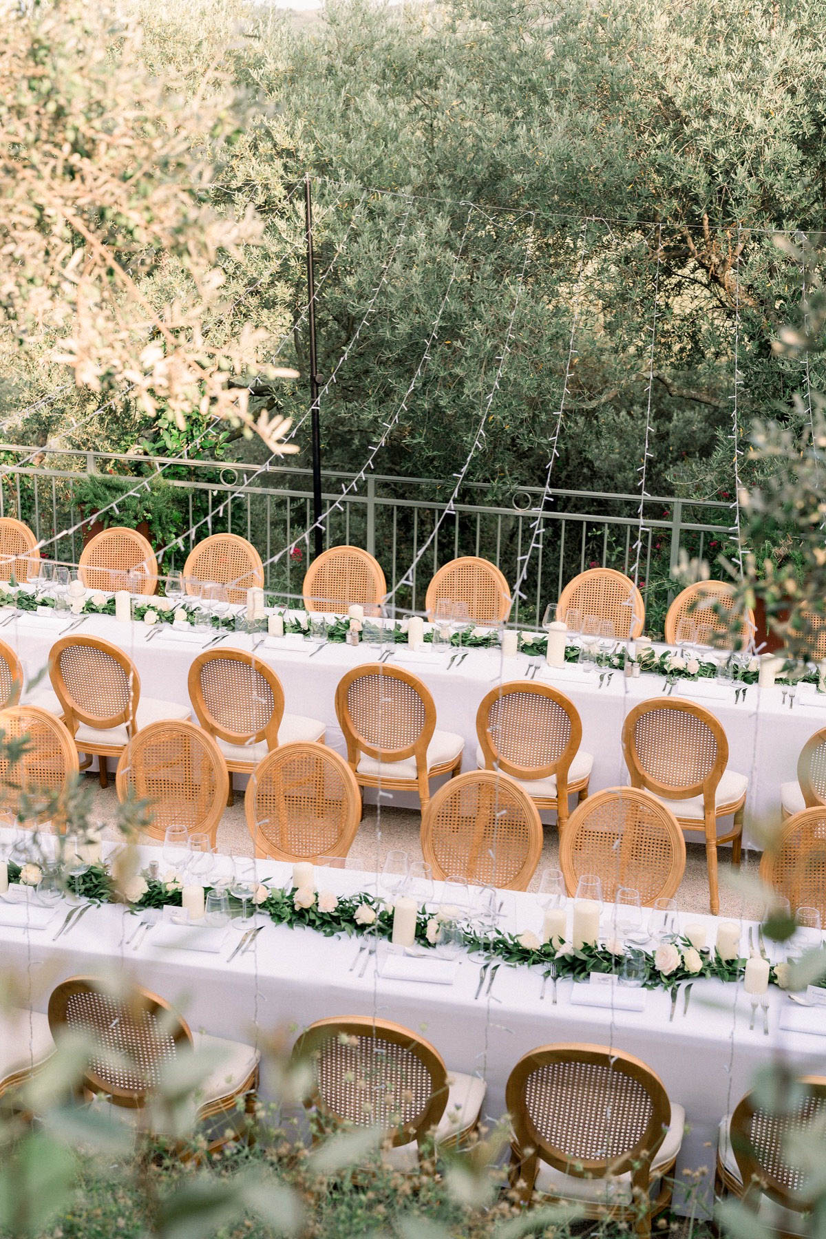 An outdoor wedding reception setup photographed from an elevated angle, showing two parallel long banquet tables dressed in white linen on a terrace. The tables are lined with natural wood-framed cane-back medallion chairs with cream upholstered seats. Table runners consist of green garlands with cream and white roses interspersed with white pillar candles, and place settings include clear glassware and silver cutlery. Fairy lights are strung overhead between poles, and a metal railing borders the terrace beyond which olive trees are visible. The overall styling is classic French with a clean white and natural wood palette.
