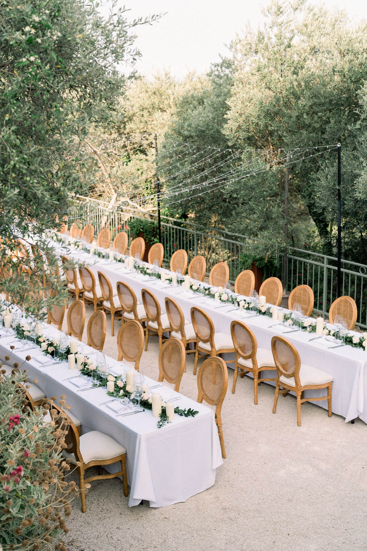 An outdoor wedding reception setup on a gravel terrace, showing two long parallel banquet tables dressed in white linen tablecloths. The tables are lined with rattan-back Louis XVI-style chairs in natural wood with cream upholstered seats. Each table features a runner of green foliage and ivory roses interspersed with white pillar candles, with glassware and place settings laid for guests. Fairy lights are strung overhead between metal poles, and a green iron railing borders the terrace edge. The overall decor palette is white, ivory, and natural wood with greenery accents, reflecting a classic French garden style. Wide overhead shot taken at a slight angle.