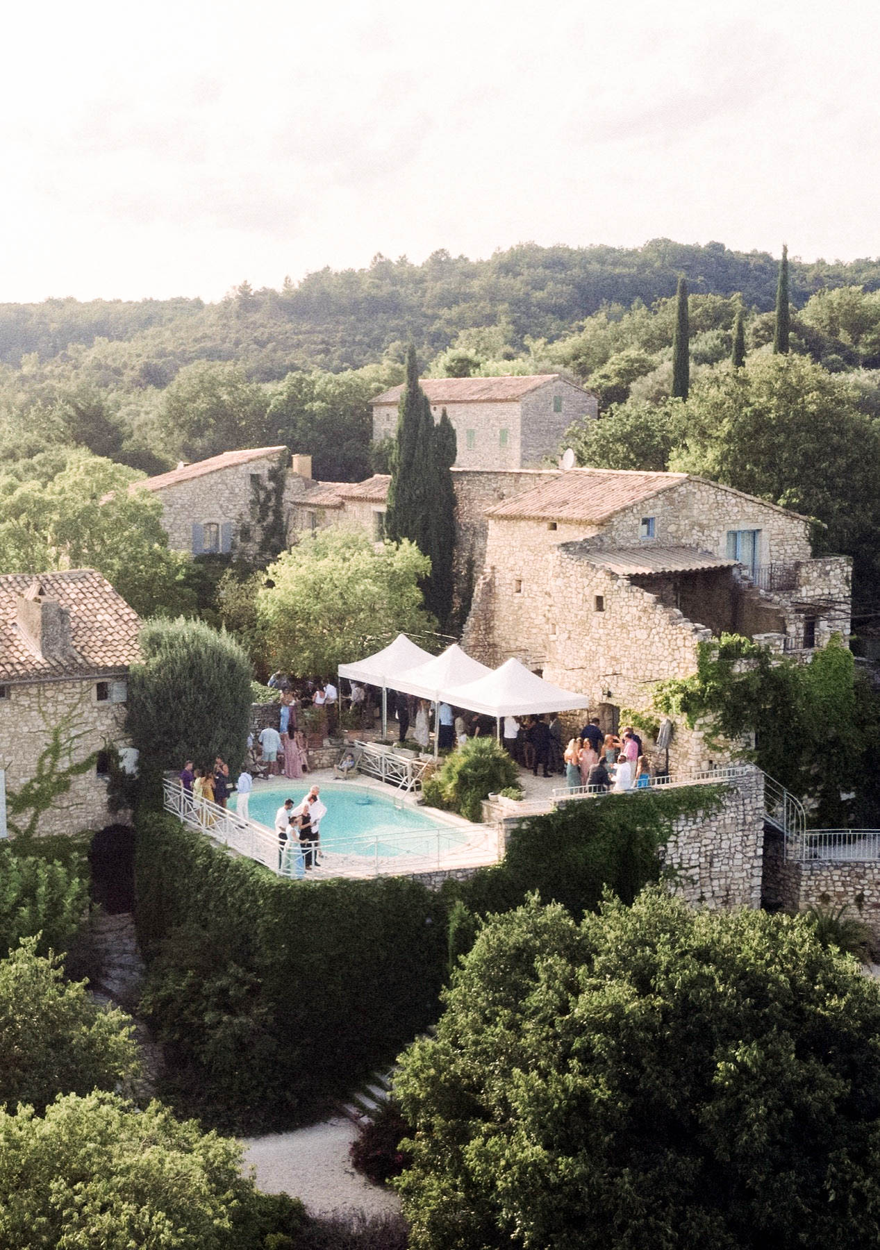 An aerial wide shot of a cocktail hour or outdoor reception taking place at a Provençal stone mas or hamlet-style property. Approximately 30–40 guests in summer wedding attire are gathered around a pool terrace and beneath two white pop-up canopy tents set up against the main stone building. A small group of guests, including what appears to be a figure in a white dress, stands near the pool edge. The venue consists of multiple interconnected stone buildings with terracotta tile roofs and blue-shuttered windows, characteristic of southern French rural architecture. The overall styling is relaxed and classic, with no heavy decor visible from this distance. Potential venue feature image.
