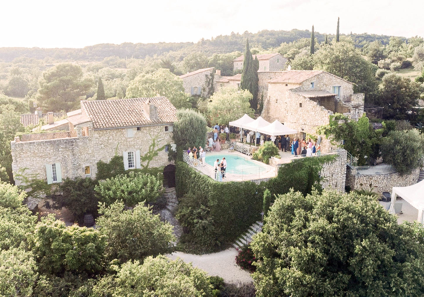 An aerial wide shot of a cocktail hour taking place at a traditional Provençal stone mas, featuring terracotta-tiled rooftops, pale blue shutters, and ivy-covered facades. Approximately 30–40 guests are gathered around a rectangular pool terrace, with two white pop-up canopy tents set up to provide shade over a reception area. Small groups of guests in colorful summer attire are mingling poolside, and what appears to be the couple stands near the pool railing. The property is composed of multiple connected stone buildings surrounded by dense Mediterranean garden foliage, with cypress trees punctuating the grounds. Potential venue feature image.